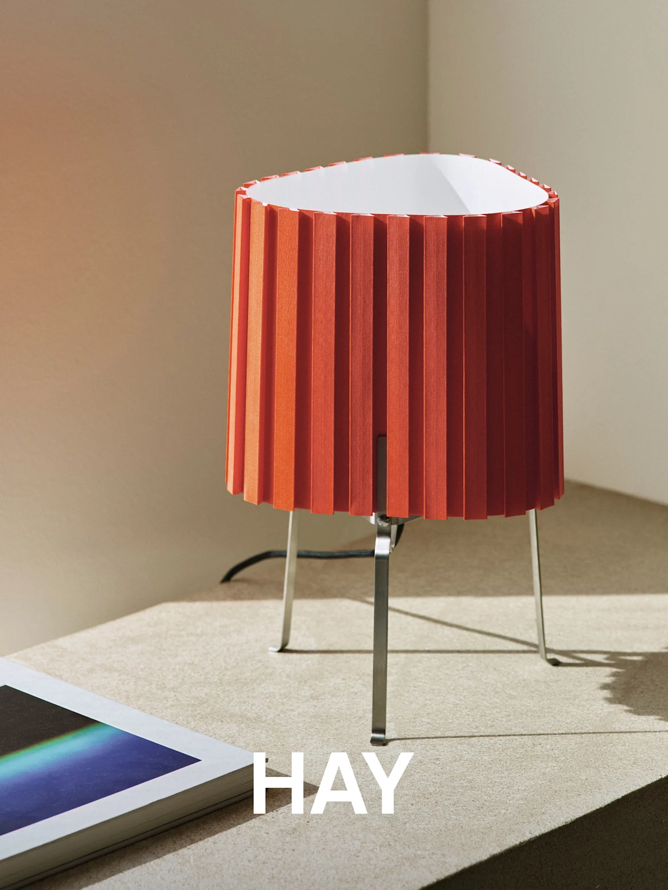 Oransje plissert bordlampe med metallfot på en teksturert overflate, ved siden av en bok. Ordet 'HAY' er overlagret.