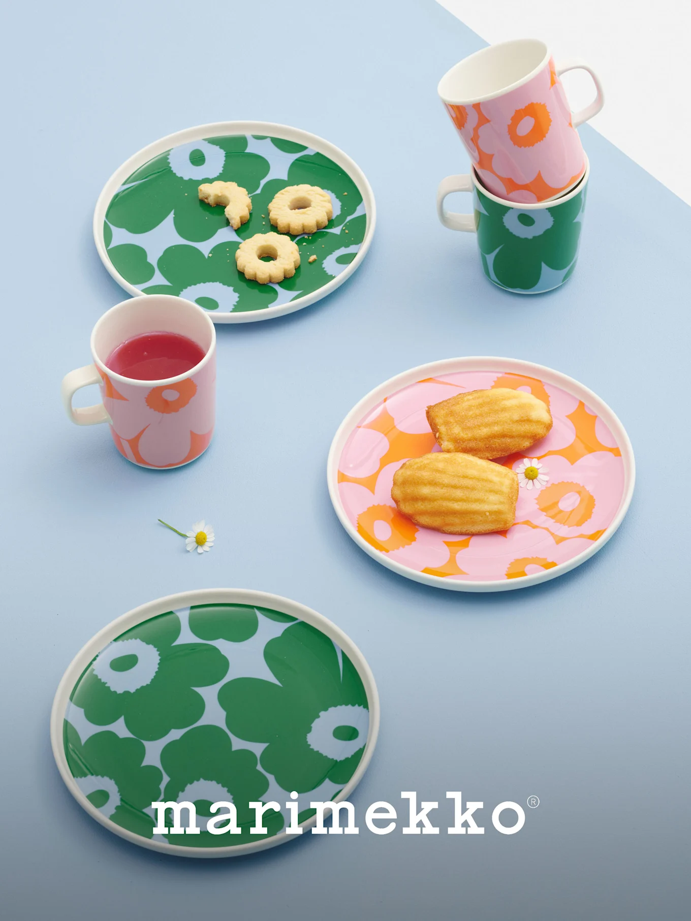 Marimekko tallerkener og krus med dristige blomstermønstre i grønt, rosa og oransje, med kjeks, madeleines og en drikke.