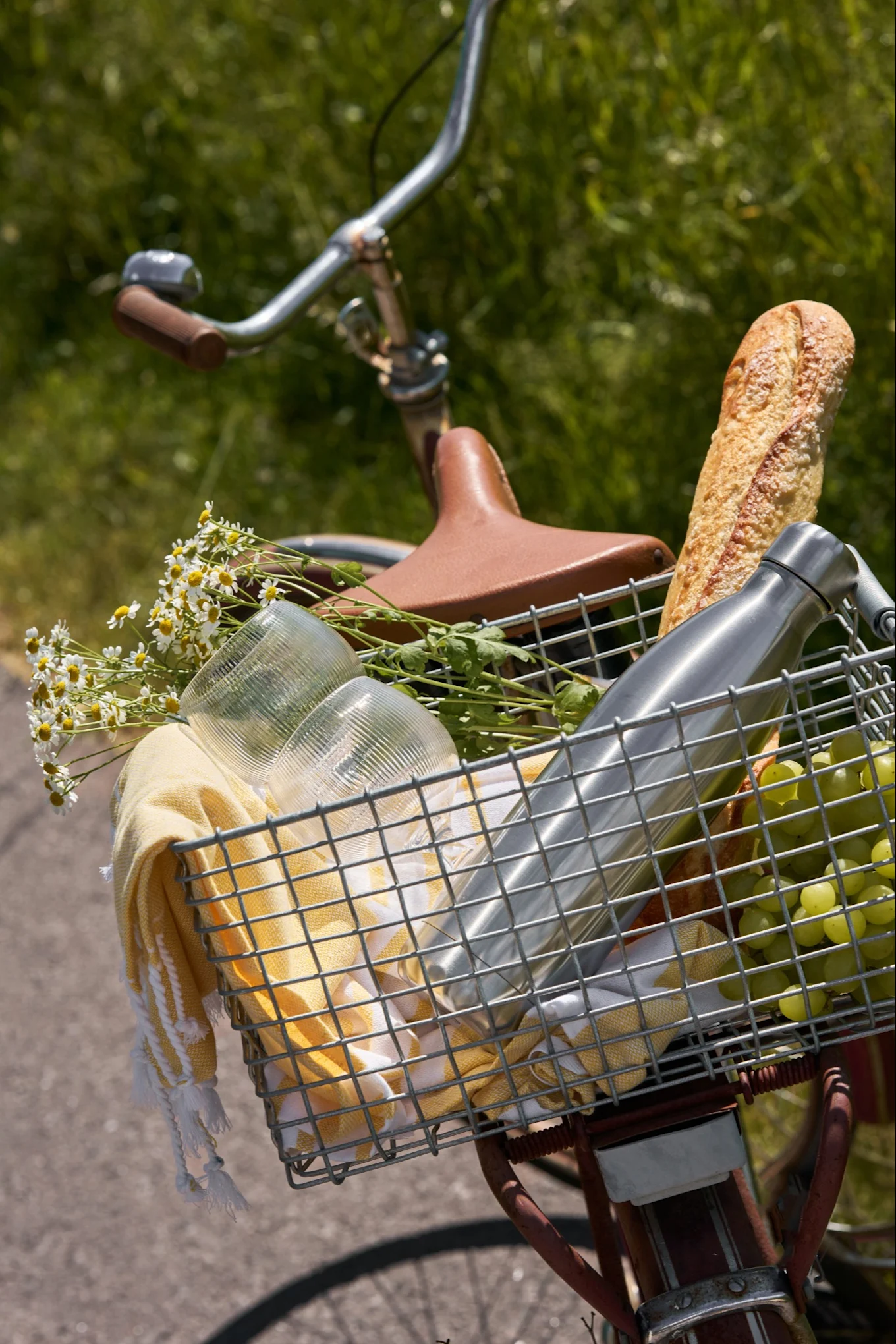 En sykkelkurv fylt med piknikutstyr: baguette, vannflaske, druer, kopper, gult håndkle og hvite tusenfryd.