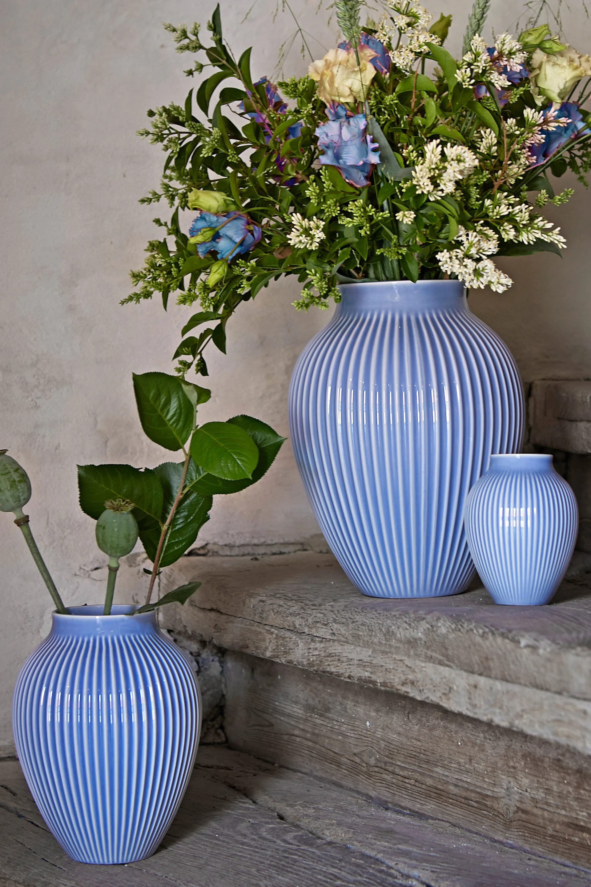 Knabstrup vase i ribbemønster fra Knabstrup keramikk står i tre forskjellige størrelser på et steintrapp, alle i samme lavendelblå farge. To av vasene har blomster i seg.