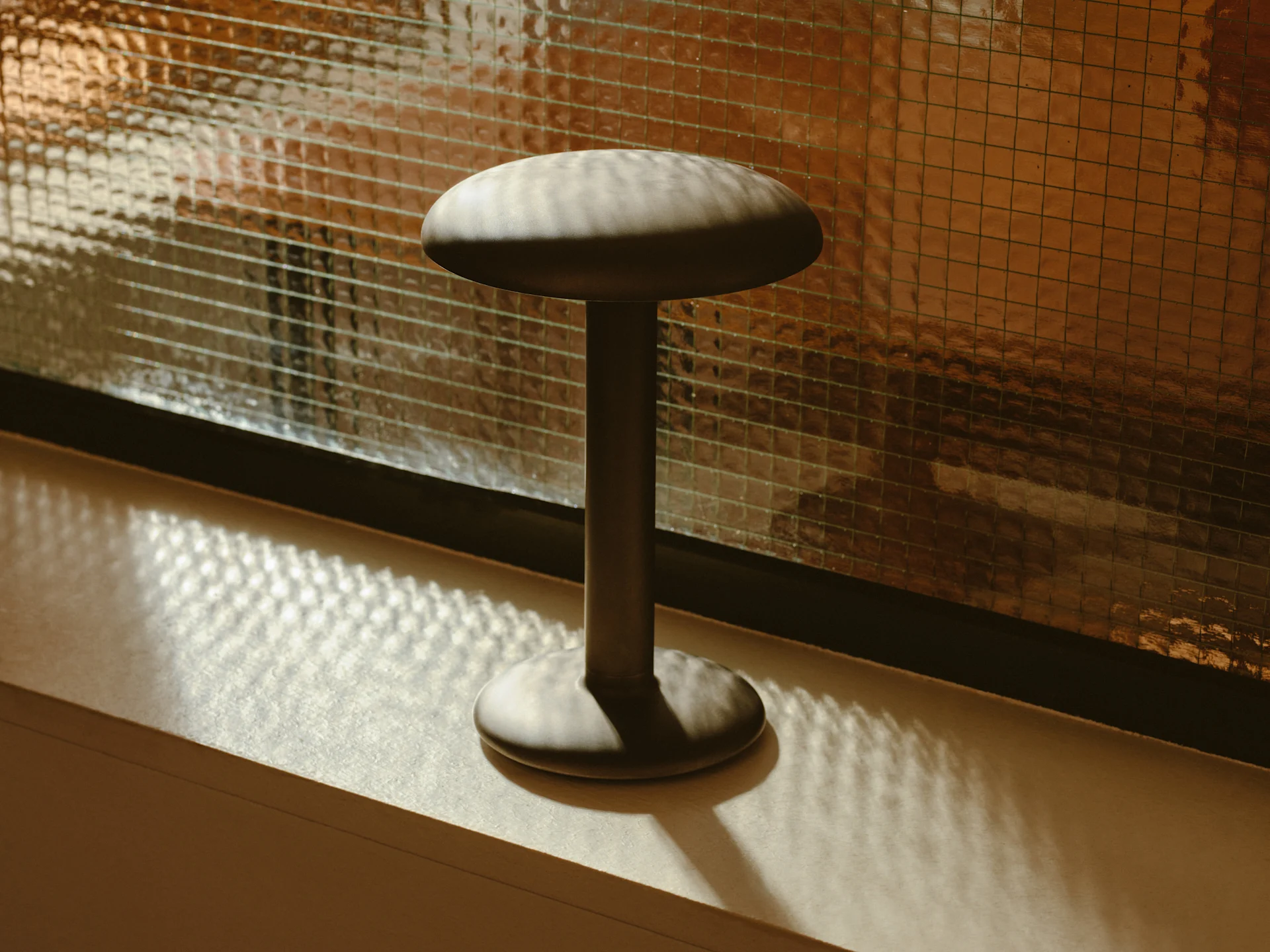 En mørk, soppformet gjenstand med flat topp og rund base står på en overflate, og kaster skygger fra et teksturert glasspanel.