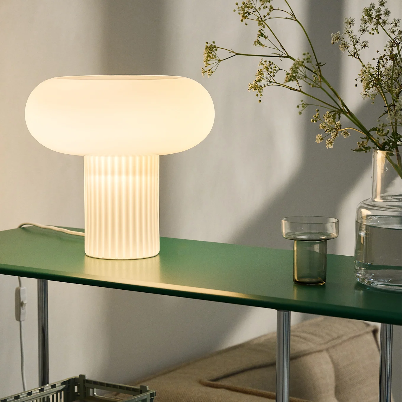 Glødende soppformet bordlampe med ribbet base på mørkegrønn hylle, sammen med blomster i en vase.
