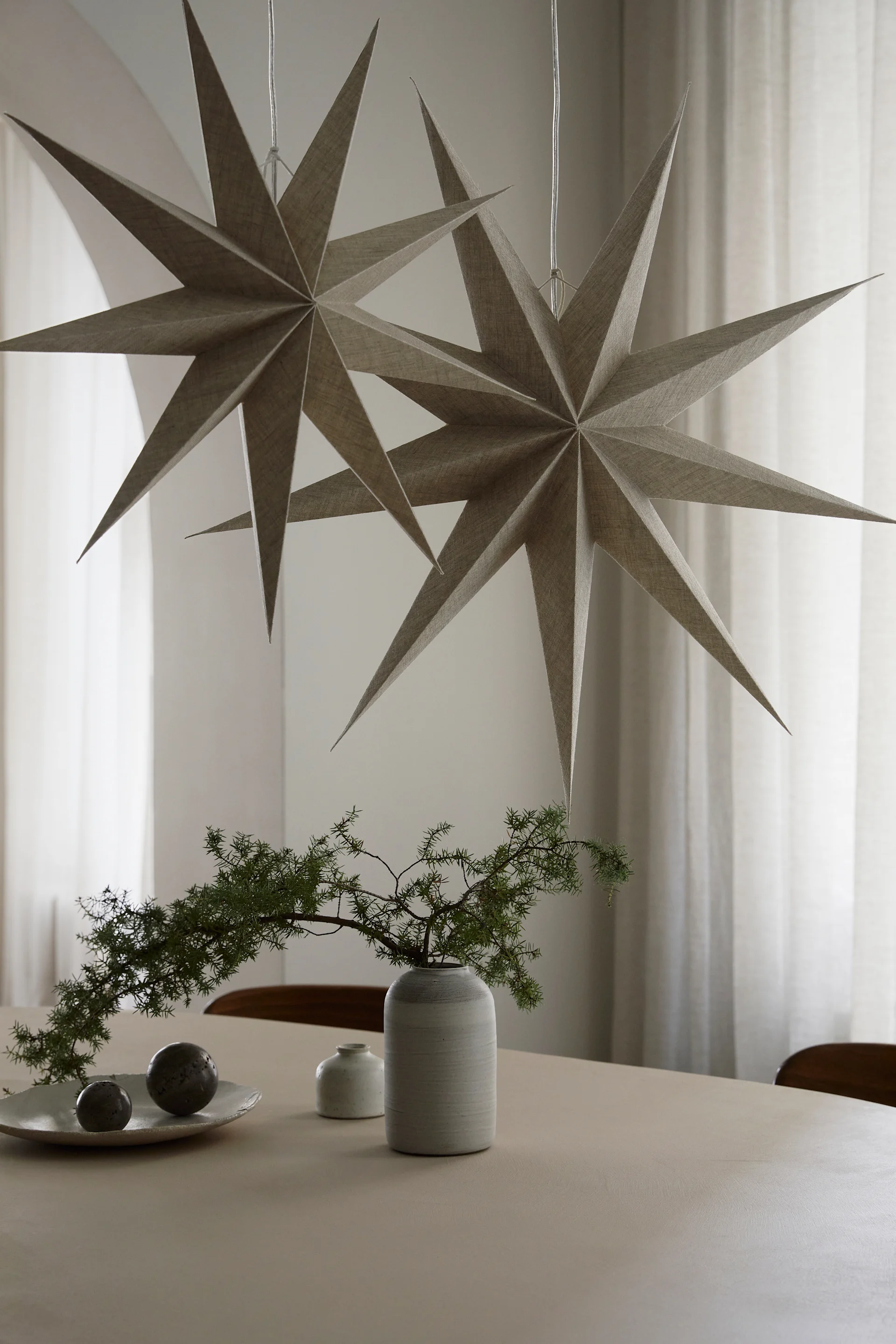 To adventsstjerner fra Watt & Veke med naturlig linfinish henger over et spisebord med en vase og noen grønne planter.
