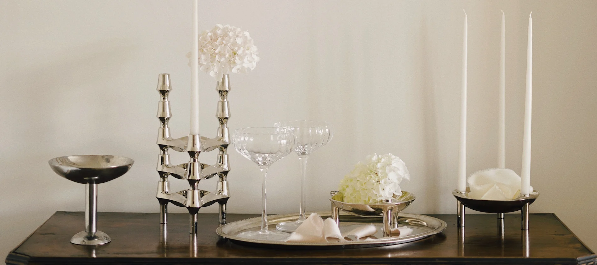 Elegant borddekorasjon med sølvlysestaker, hvite lys, glass og hvite hortensia på treflate.
