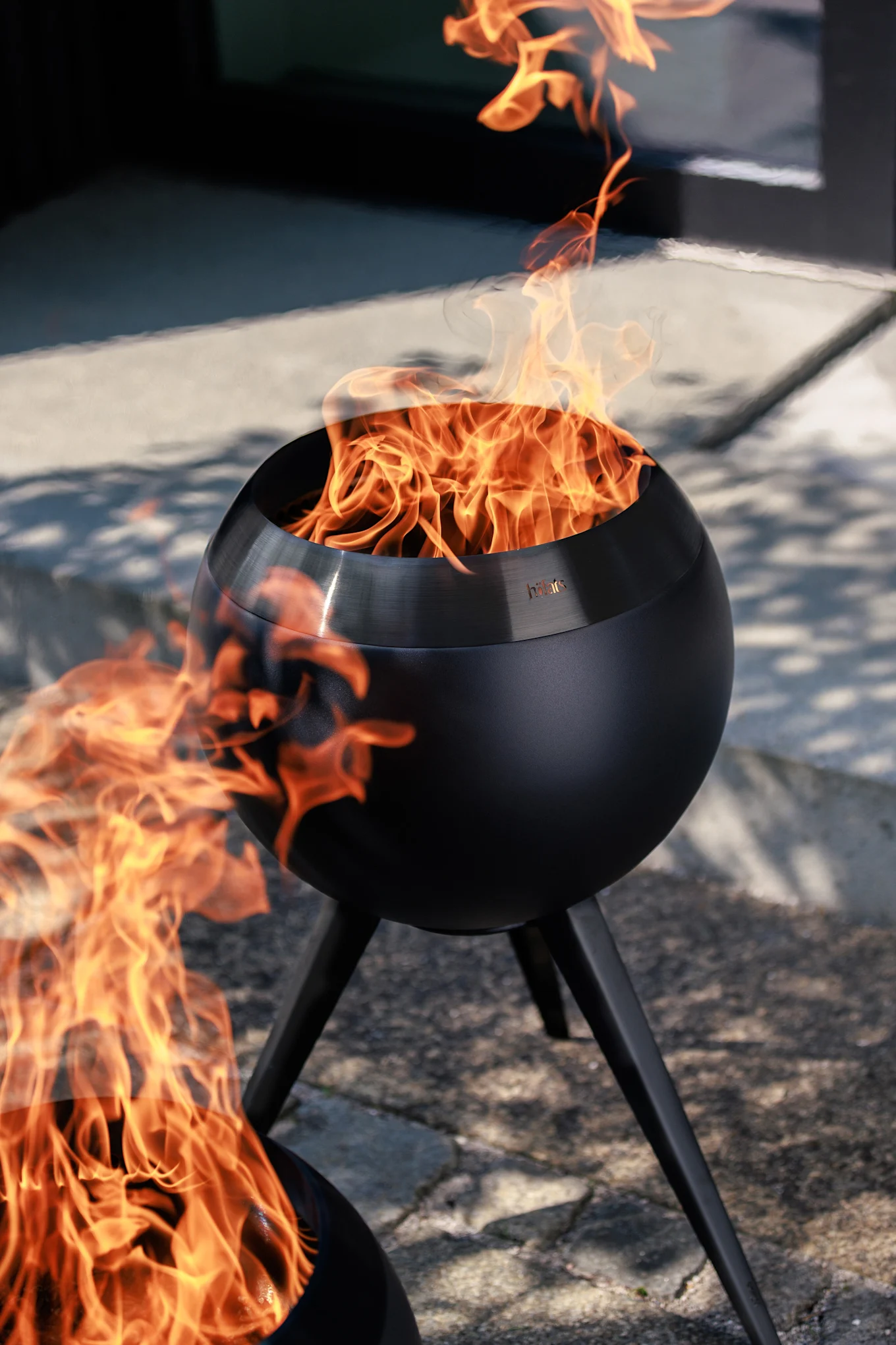 To svarte, kuleformede utendørs bålpanner med lyse oransje flammer på en brolagt uteplass.