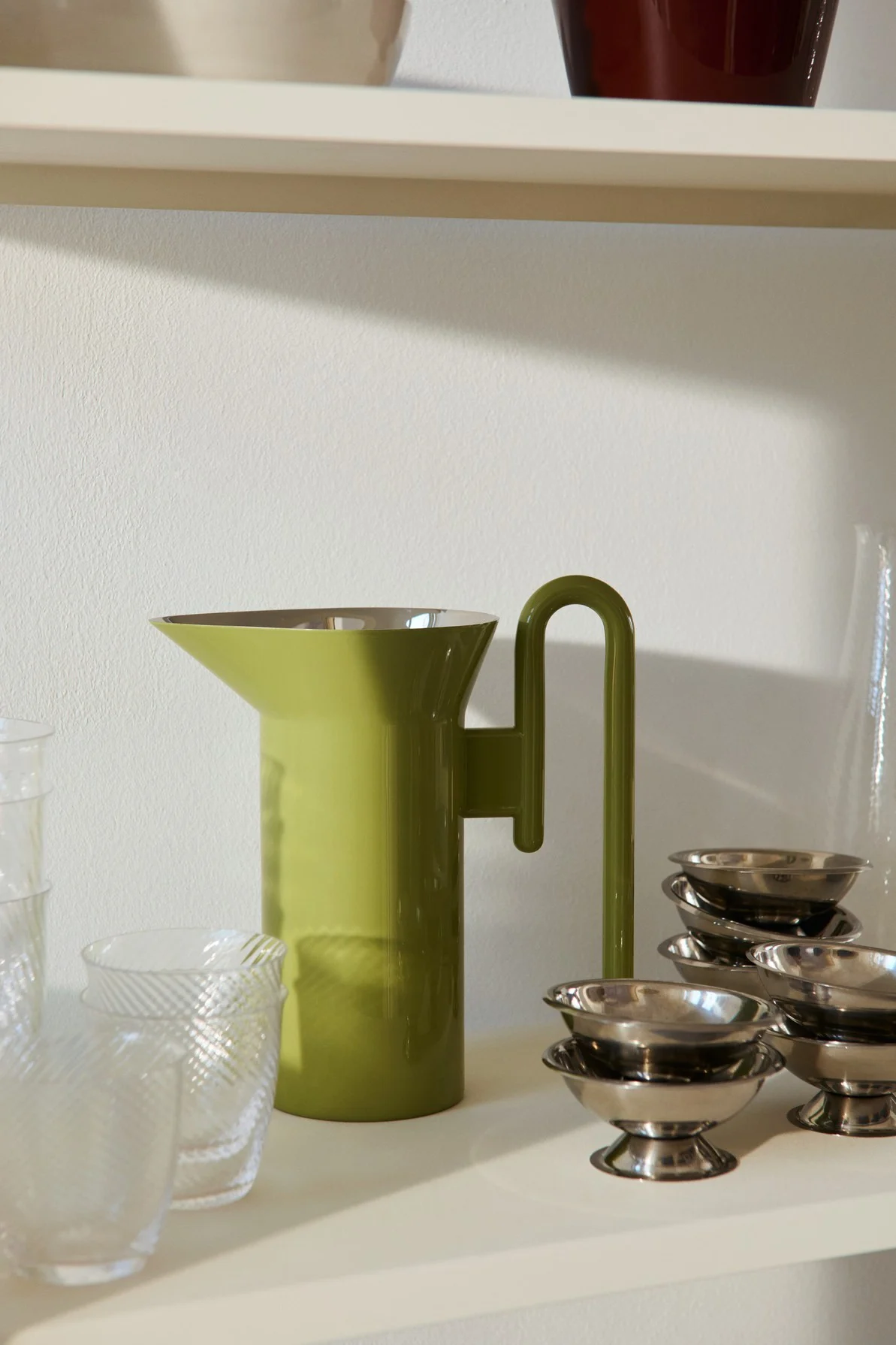 Olivengrønn mugge med U-formet håndtak, flankert av klare mønstrede glass og stabler med små sølv dessertskåler på en hylle.