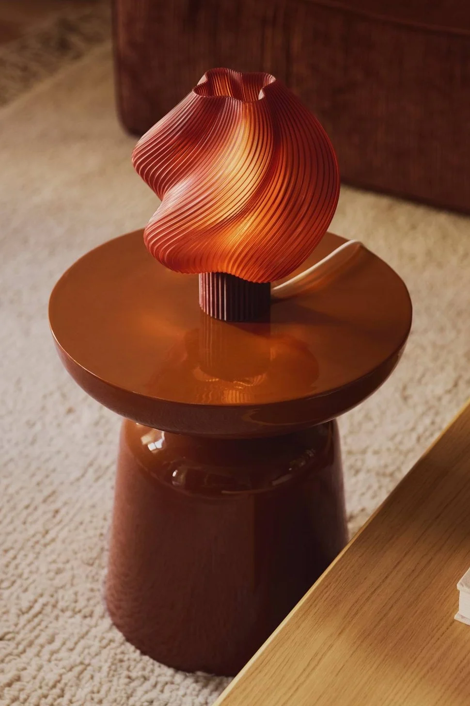 En varmt opplyst, bølget oransje plissert lampe står på et blankt brunt sidebord.