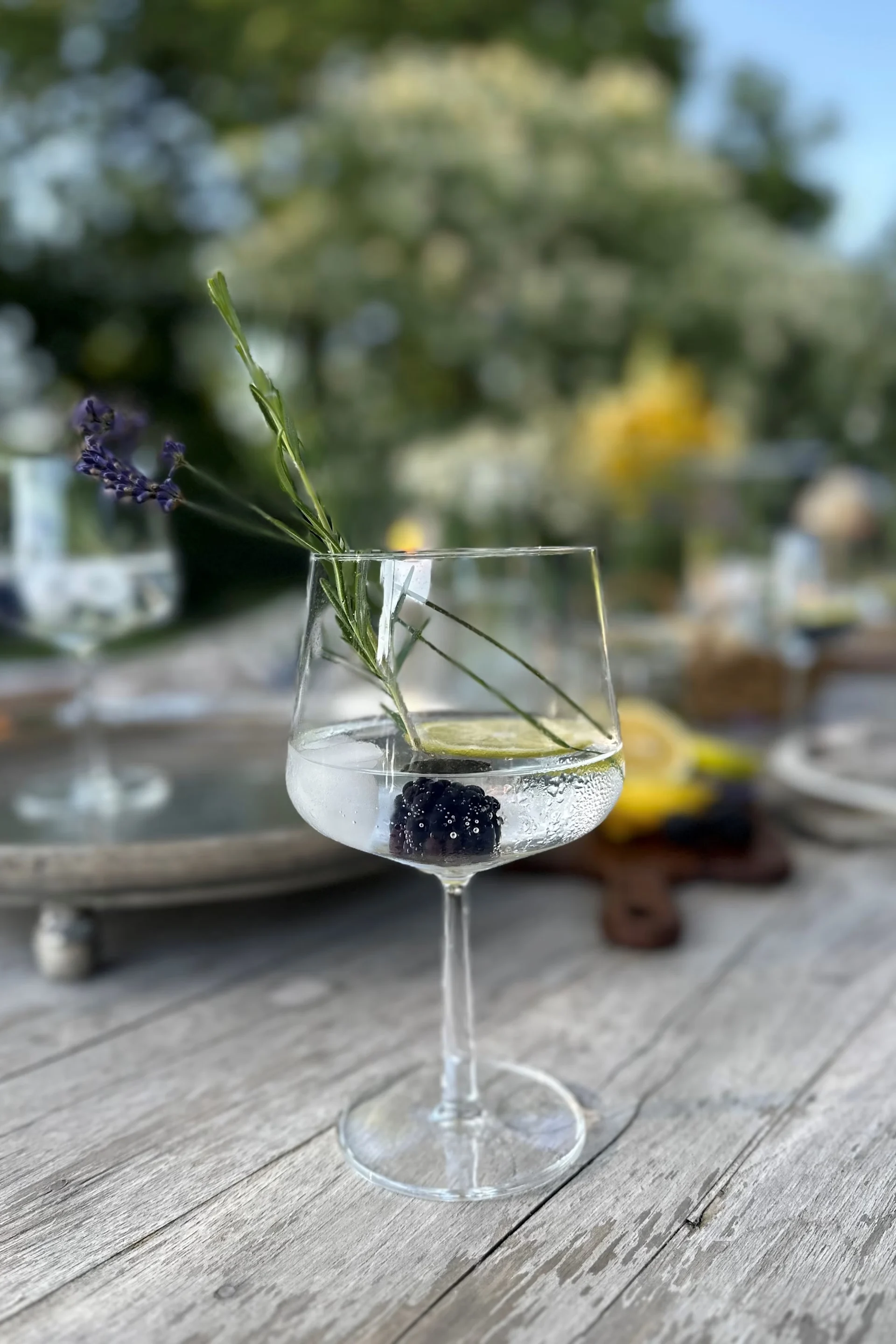 En stilkglasscocktail med et bjørnebær, limeskive, lavendel og en grønn urt på et trebord.
