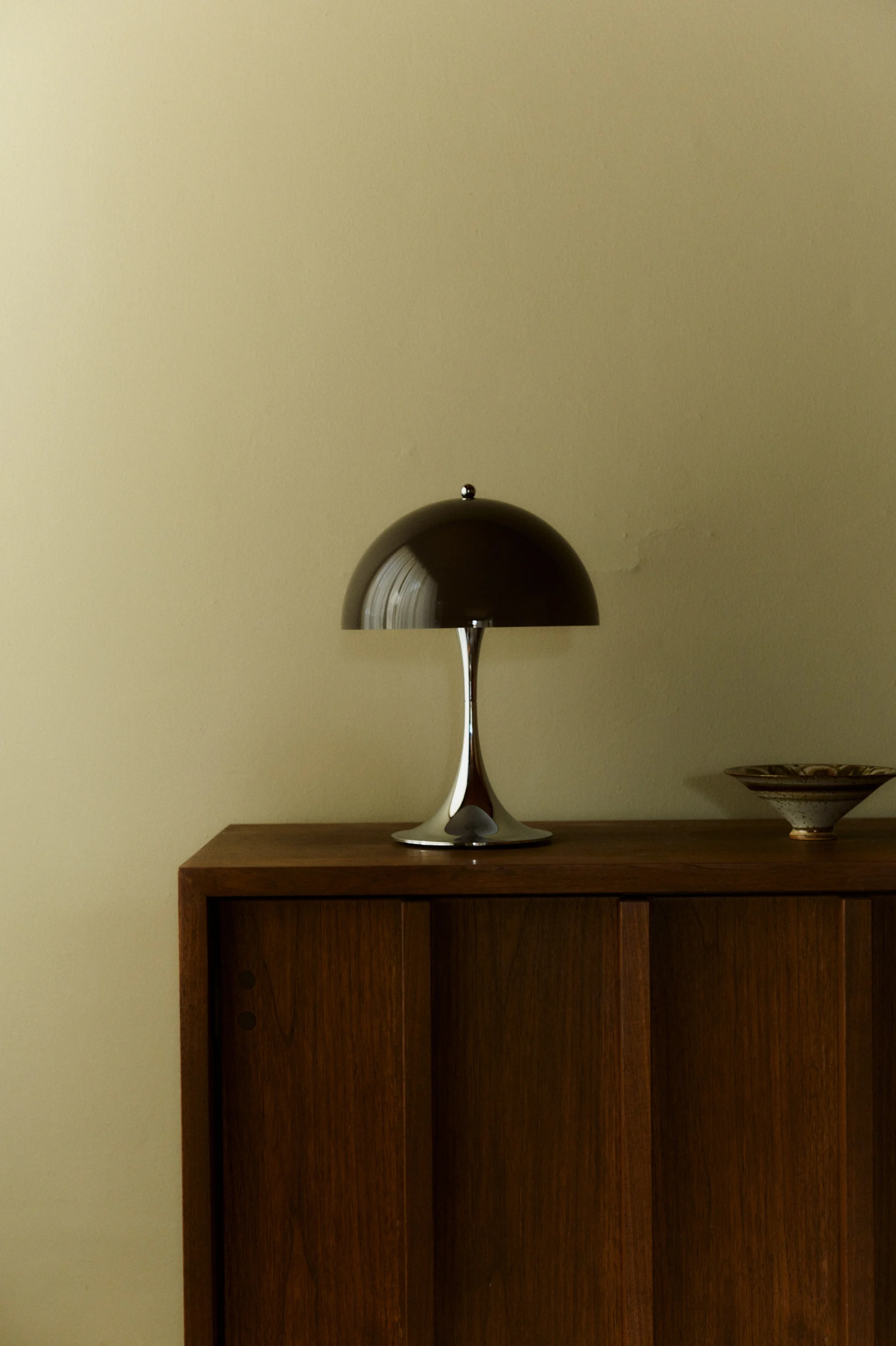 En elegant, moderne soppbordlampe med mørk kuppelformet skjerm og kromfot står på et treskap ved siden av en keramisk bolle.
