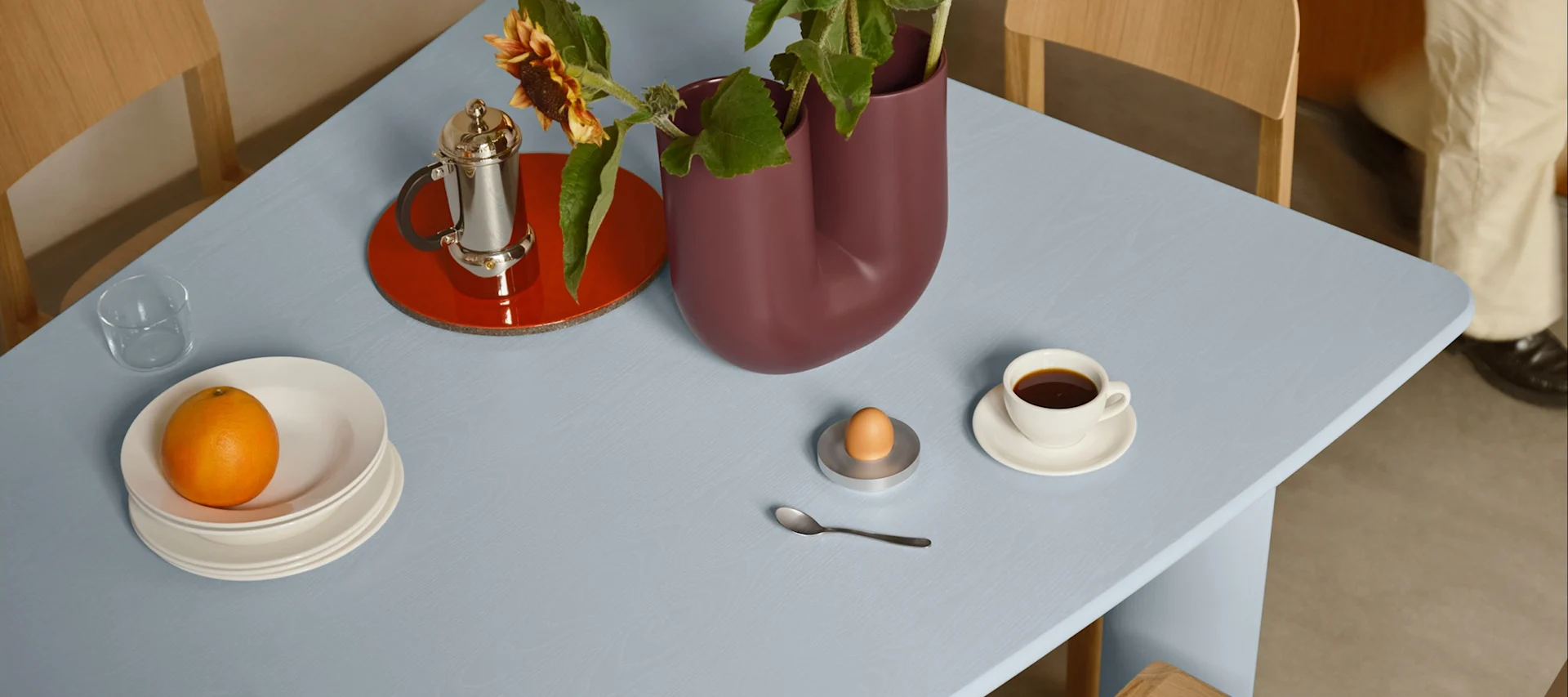 Lyseblått bord dekket med frokost: appelsin på tallerkener, kaffe, egg i eggeglass og en vase med solsikker.