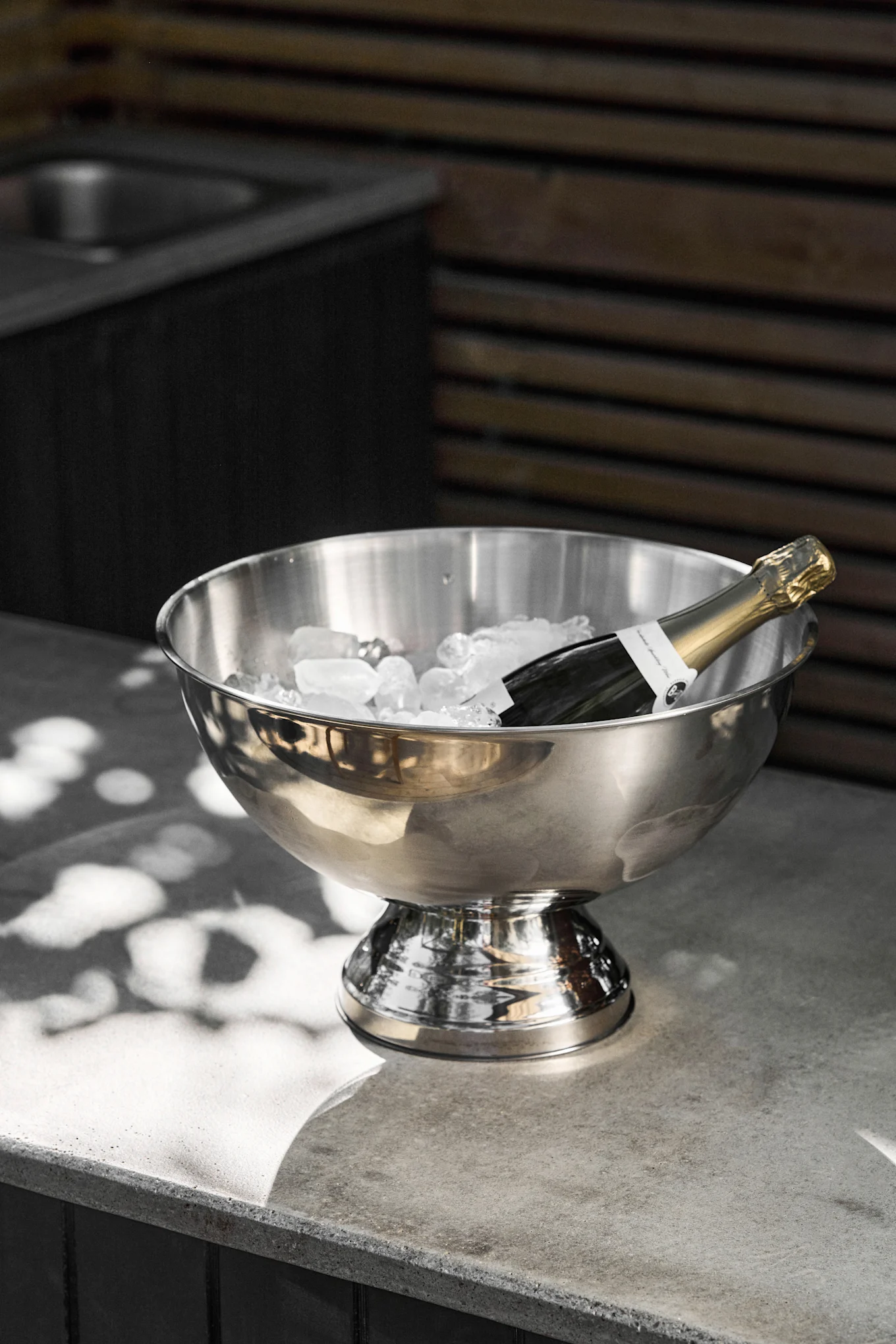 En champagneflaske ligger i en isbøtte i rustfritt stål fylt med isbiter på en grå betongbenk, med flekkvis sollys.