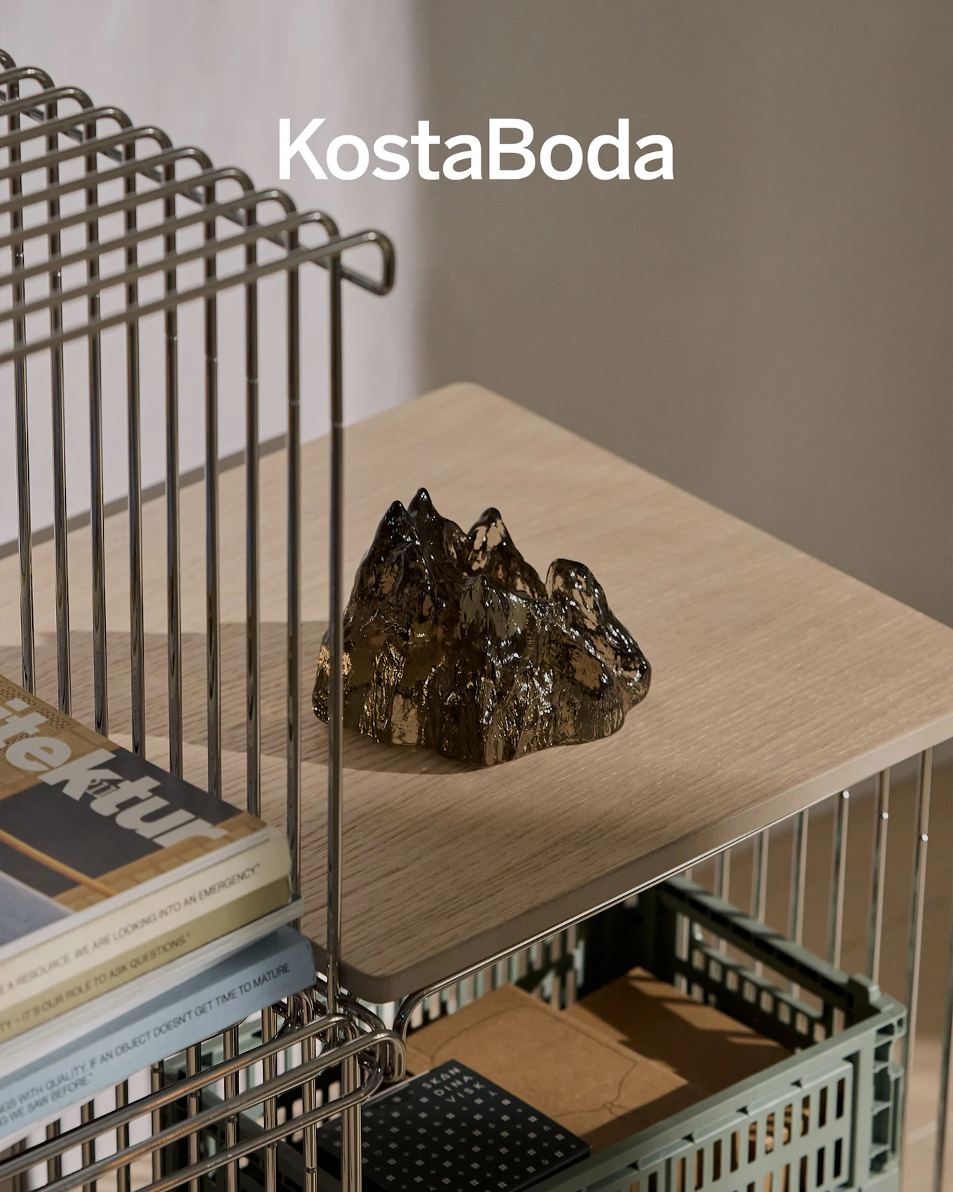 KostaBoda-merkenavn over en mørk, transparent, fjellformet glasskulptur på en lys trehylle.