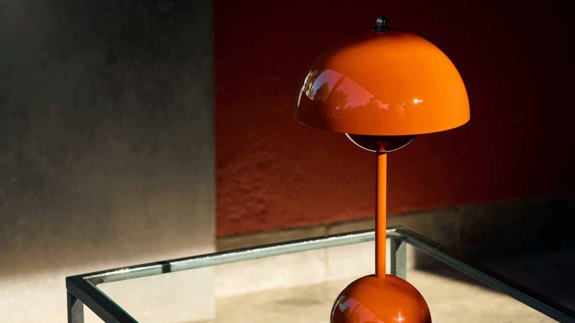 Blank, oransje soppformet bordlampe på et glassbord mot en rød og grå vegg, med flekkete sollys.