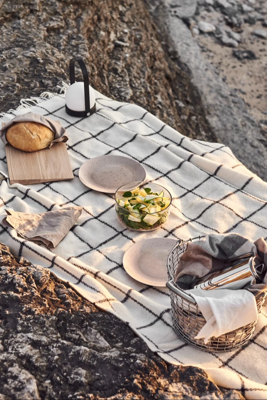 Piknik på steiner med rutet teppe, brød, lanterne, salat, tallerkener og kurv.