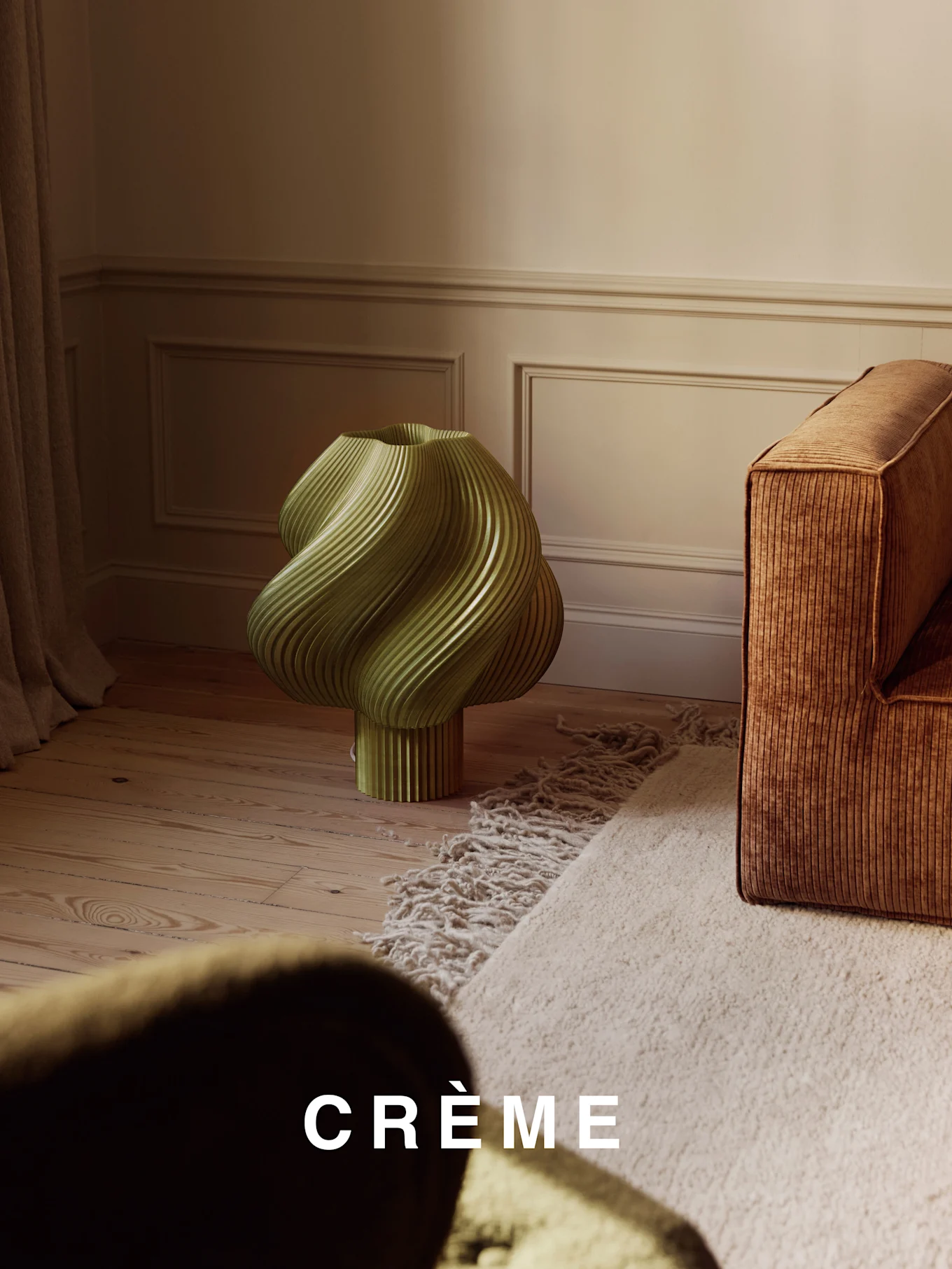 En lysegrønn, vridd ribbet lampe på et tregulv, ved siden av en brun ribbet sofa og et beige raggete teppe.