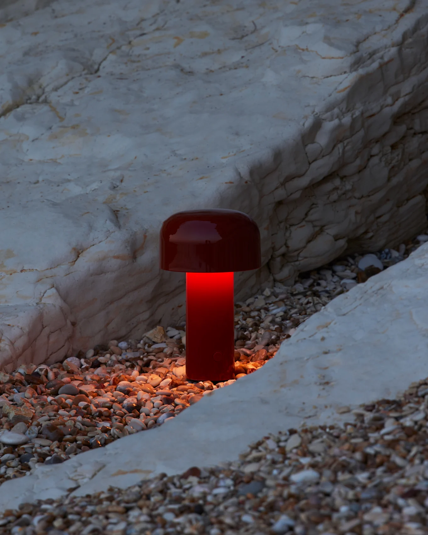 En rød soppformet lampe lyser opp en rullesteinstrand med en varm oransje glød mot lyse steiner.