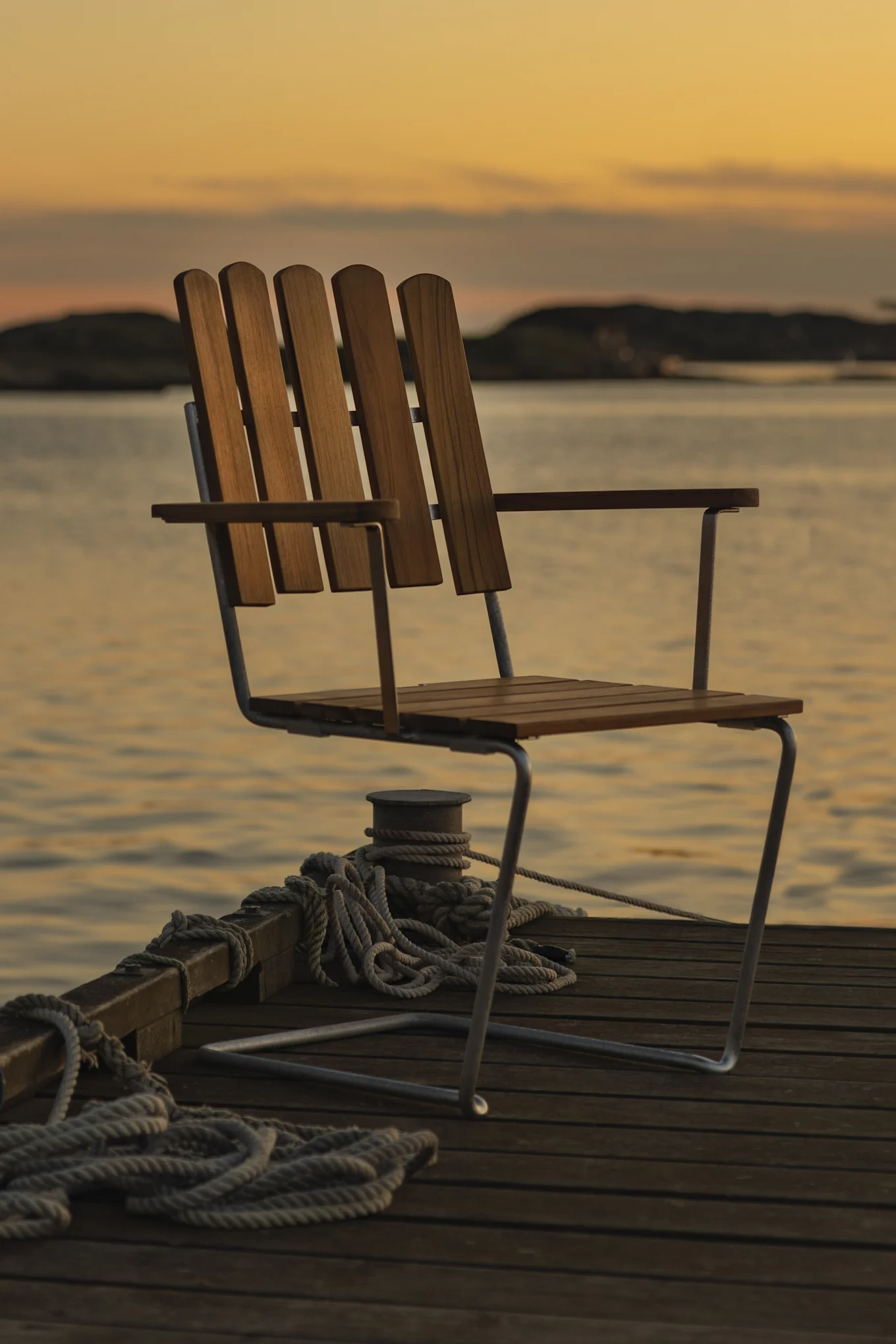 Tre stol på en brygge ved solnedgang med vann og svake landområder i bakgrunnen.