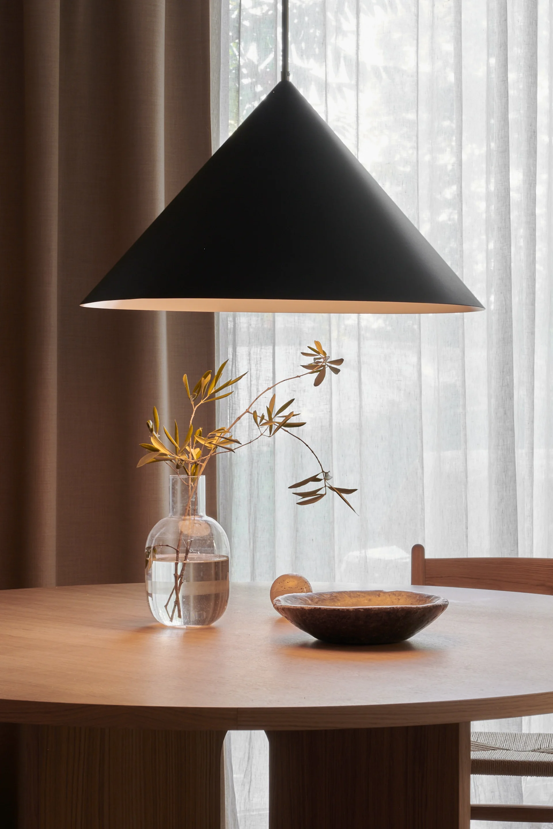 Rundt spisebord i tre med svart konisk pendellampe over, med en klar vase med grener og en mørk treskål.