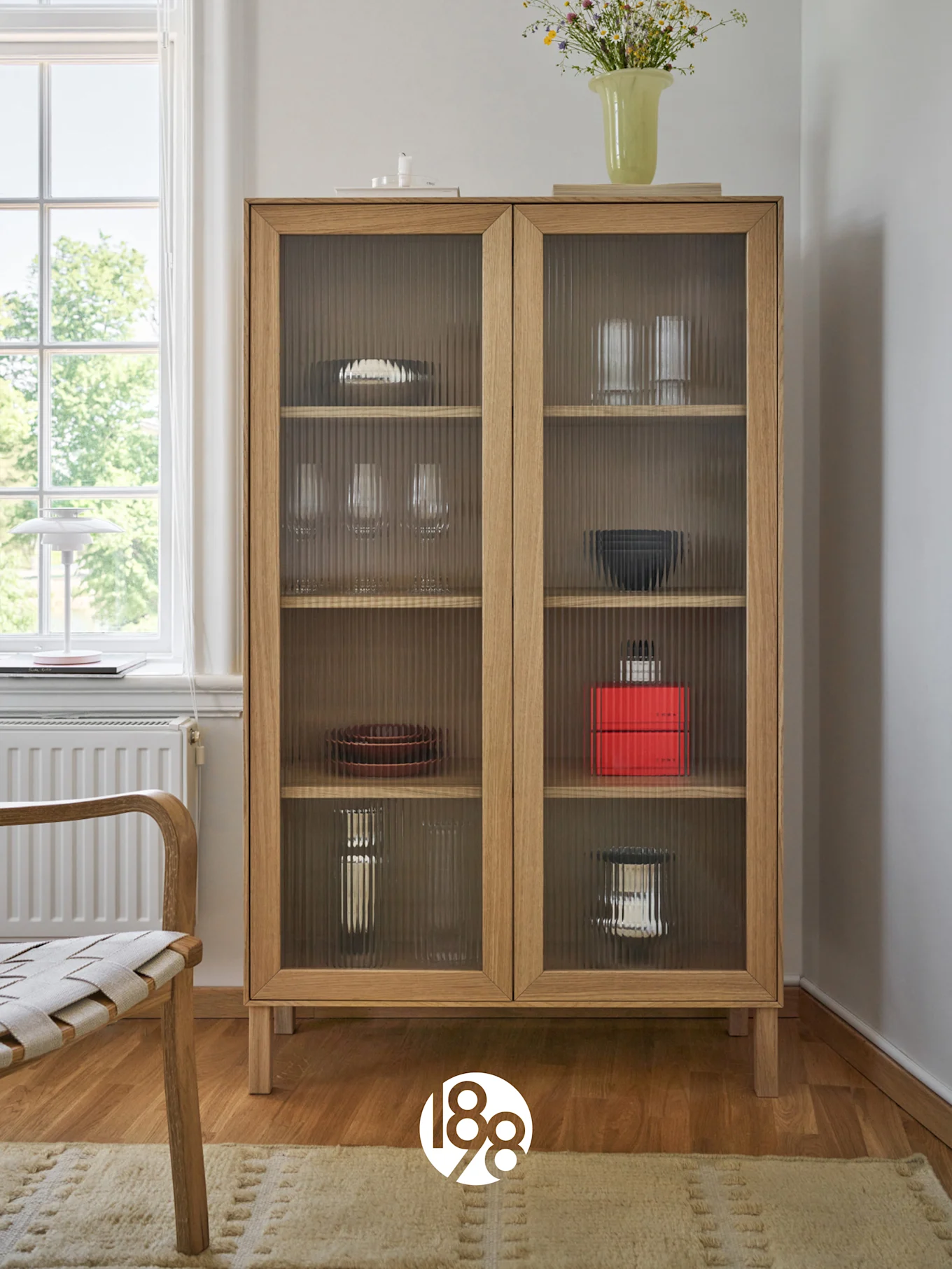 Et lyst treskap med riflede glassdører som viser servise og glass, toppet med en vase med villblomster. En stol og et vindu er til venstre.