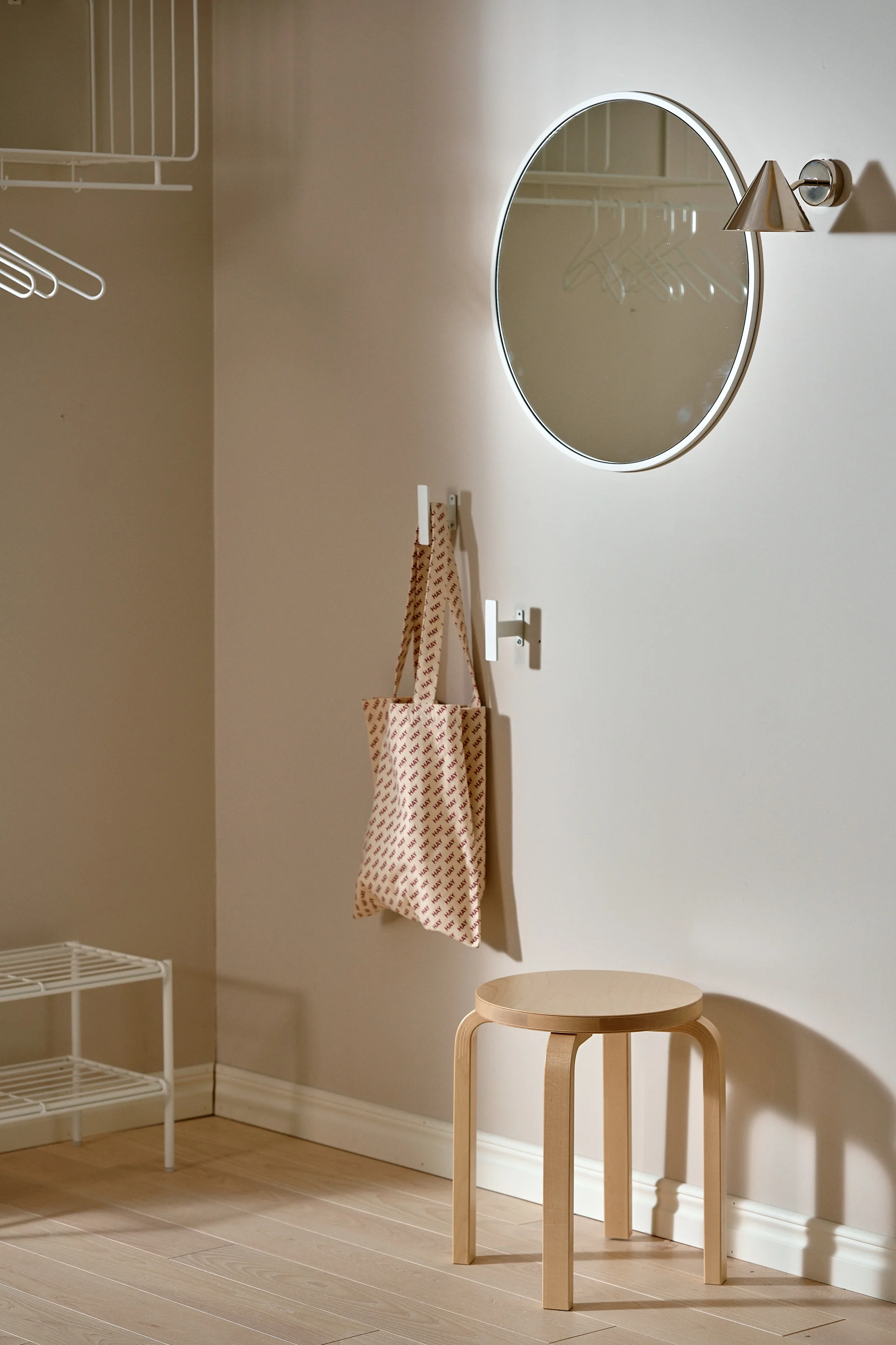 En minimalistisk inngang med et rundt speil, en trekrakk, en 'HAY'-mønstret handlenett på en krok og hvite trådhyller.