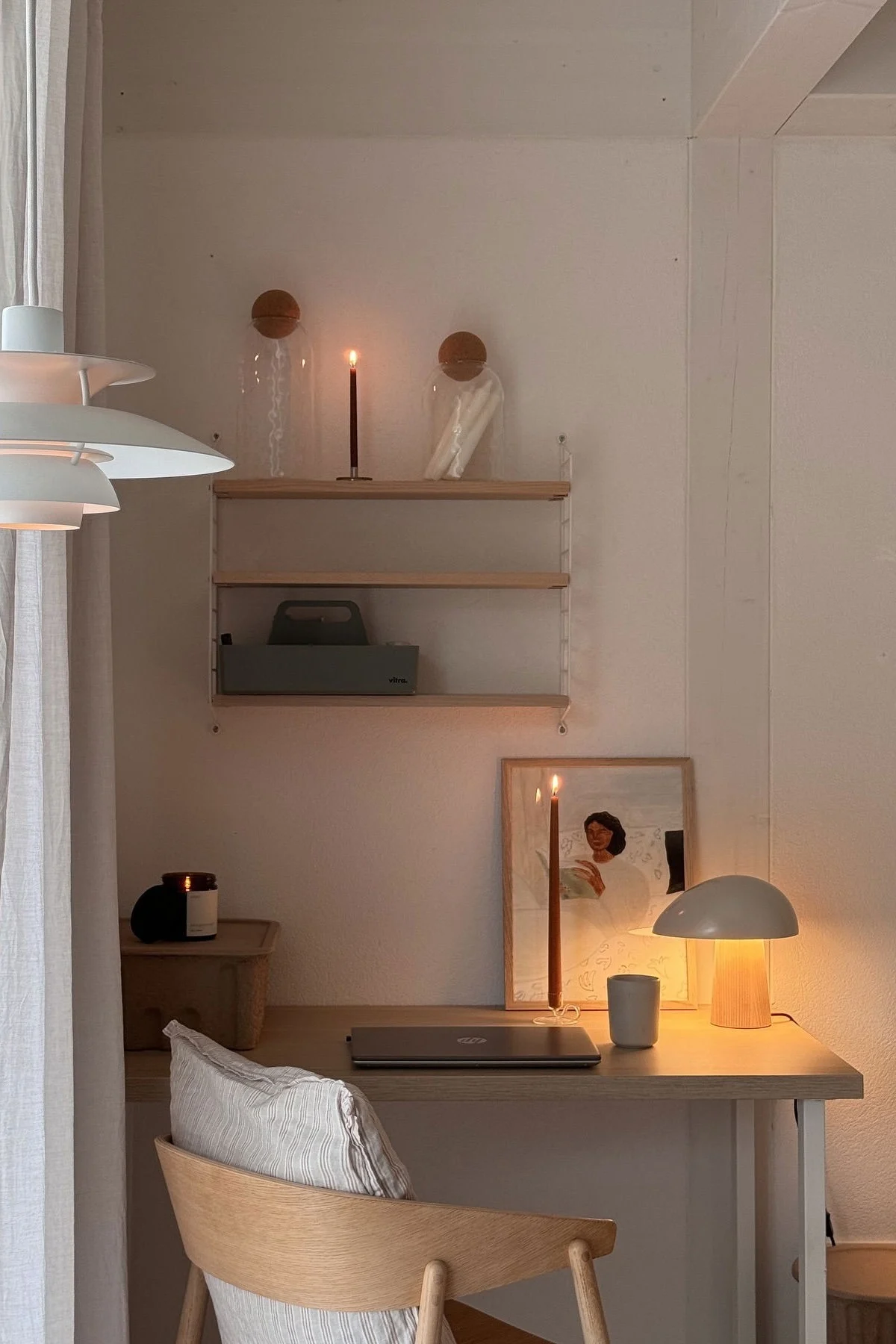 Koselig hjemmekontor med hvit pendellampe, skrivebord i tre, bærbar datamaskin, tente stearinlys og en varm soppbordlampe.