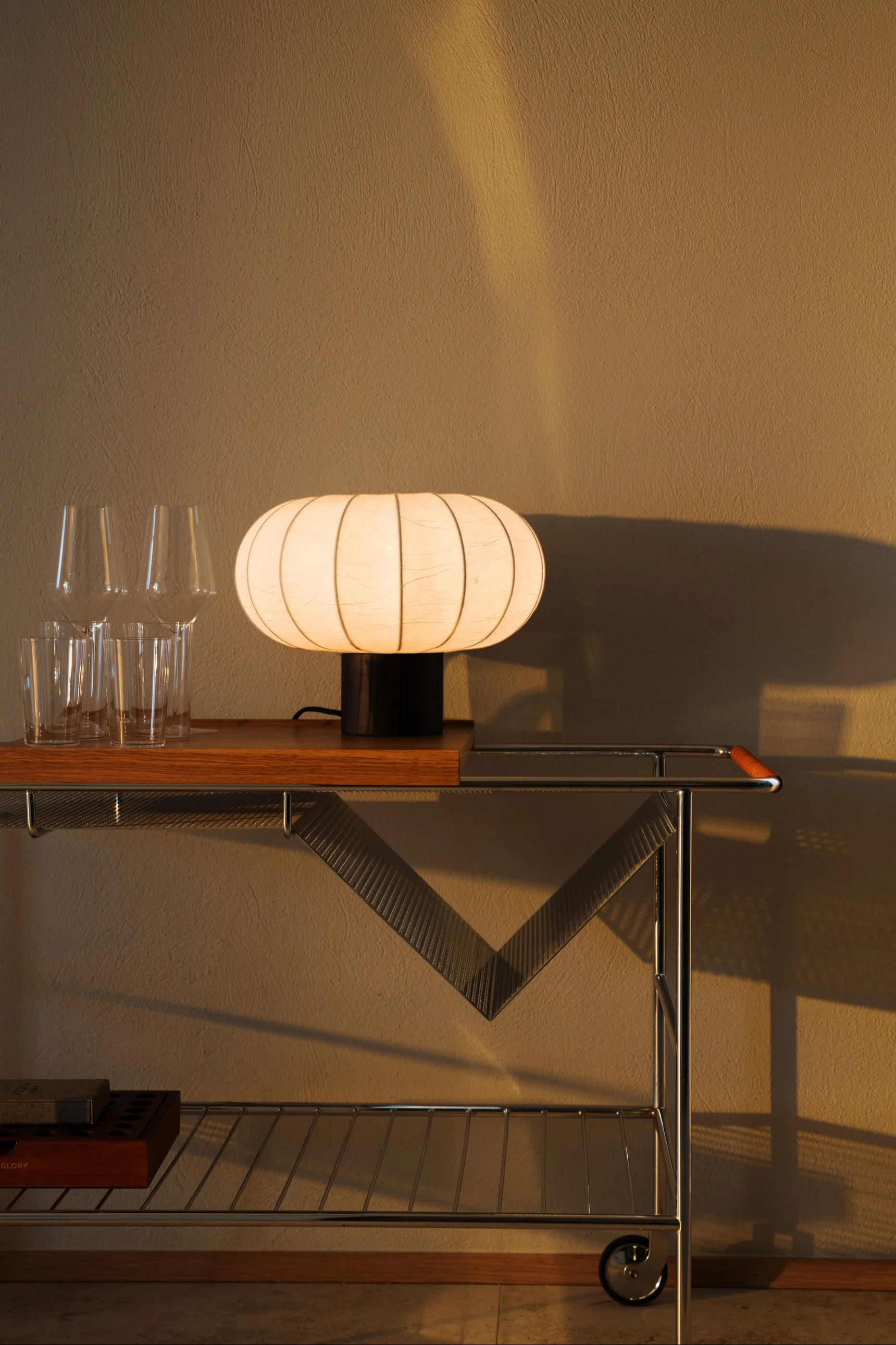 En glødende, ribbet, oval lampe står på en barvogn av tre og krom ved siden av klare glass, med en teksturert vegg i bakgrunnen.