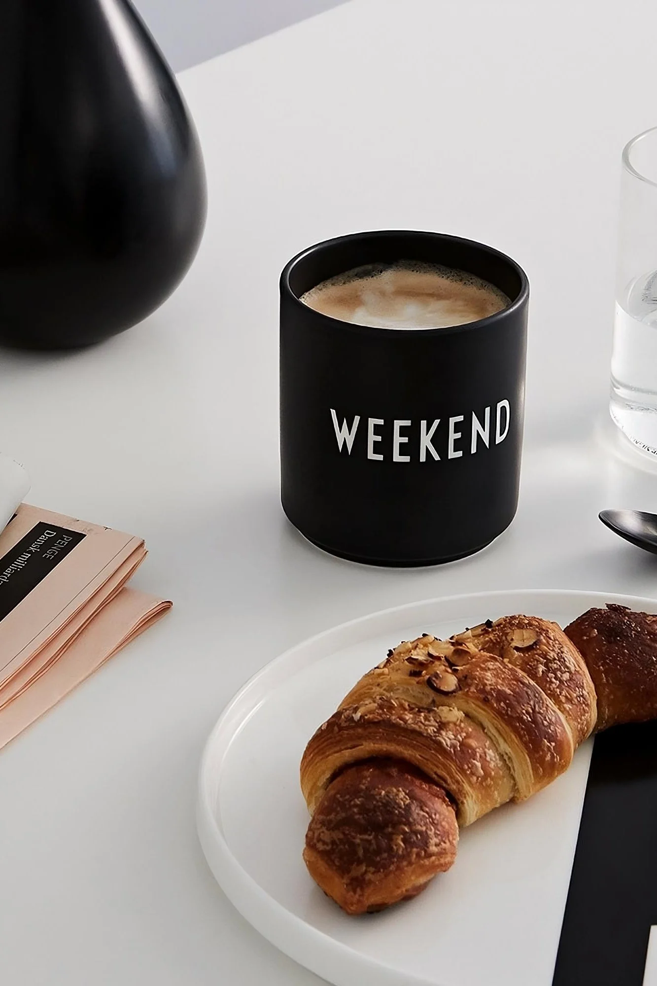 Kaffekopp: Den svarte kaffekoppen med teksten «Weekend» fra Design Letters står på et bord, foran den ligger en tallerken med en croissant.
