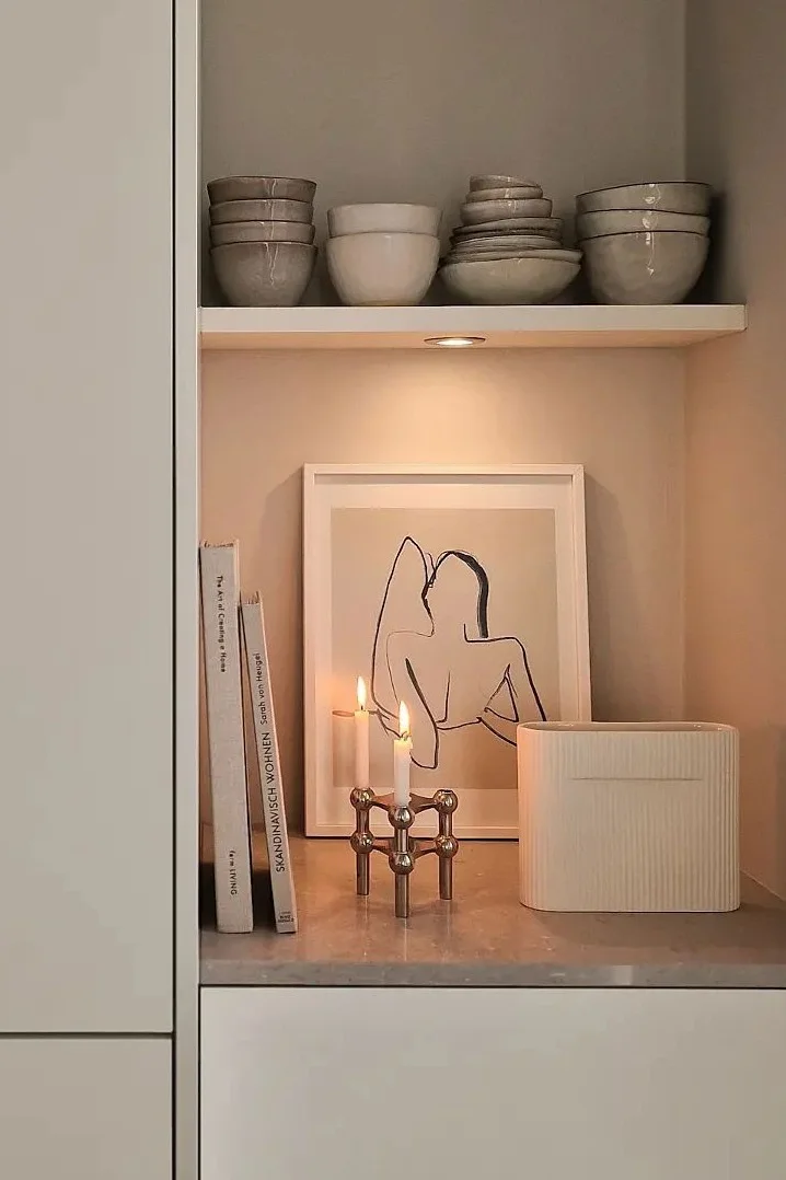 En koselig hylle med keramikkskåler, bøker, innrammet kunst, tente lys i en messingholder og en riflet vase.