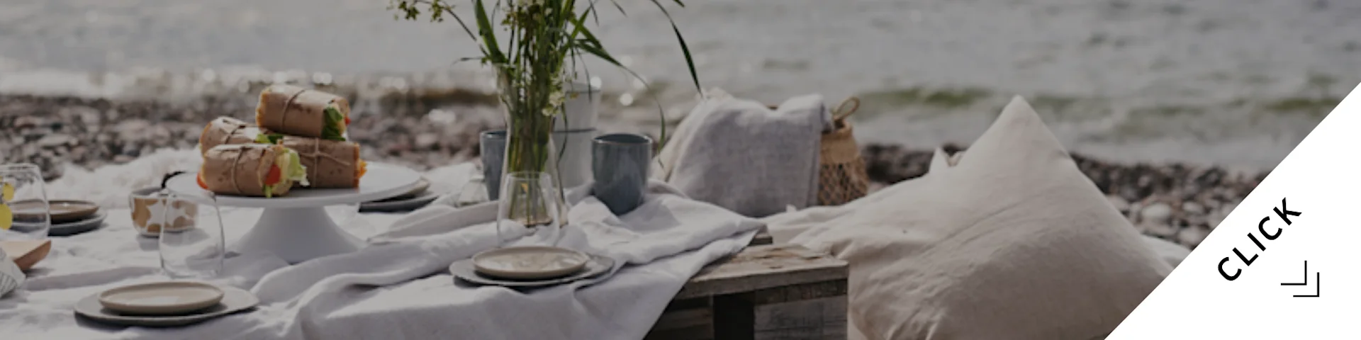 En rolig strandpiknik med innpakket mat, servise, en vase med blomster og puter på en rullesteinstrand ved sjøen.