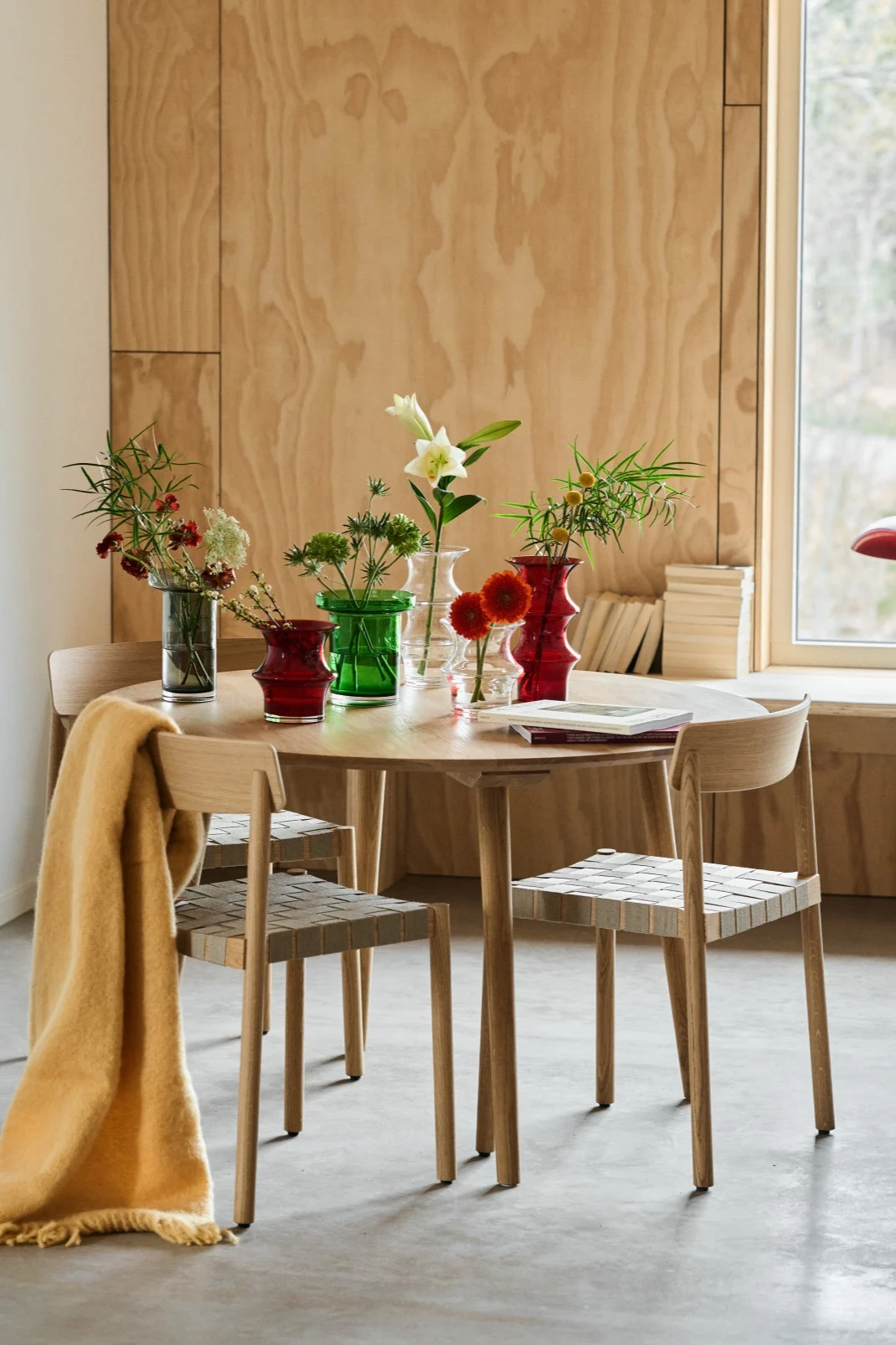 Et spisebord midt i et kryssfinerrom med en samling røde, grønne, grå og klare vaser.