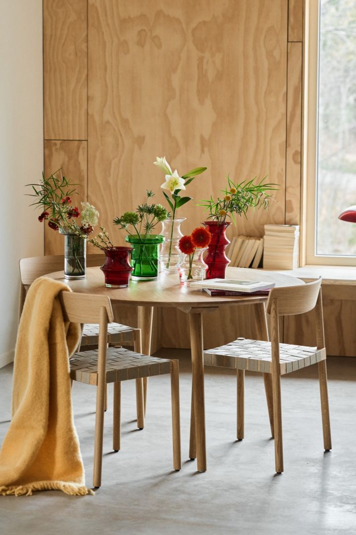 Et spisebord midt i et kryssfinerrom med en samling røde, grønne, grå og klare vaser.