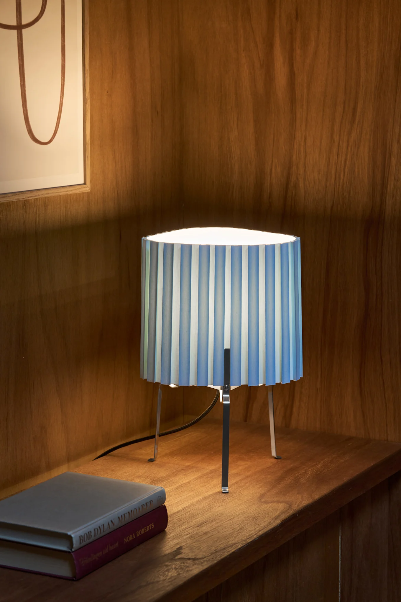 En bordlampe med blå- og hvitstripet plissert skjerm og metallben, stående på en trehylle ved siden av to bøker.