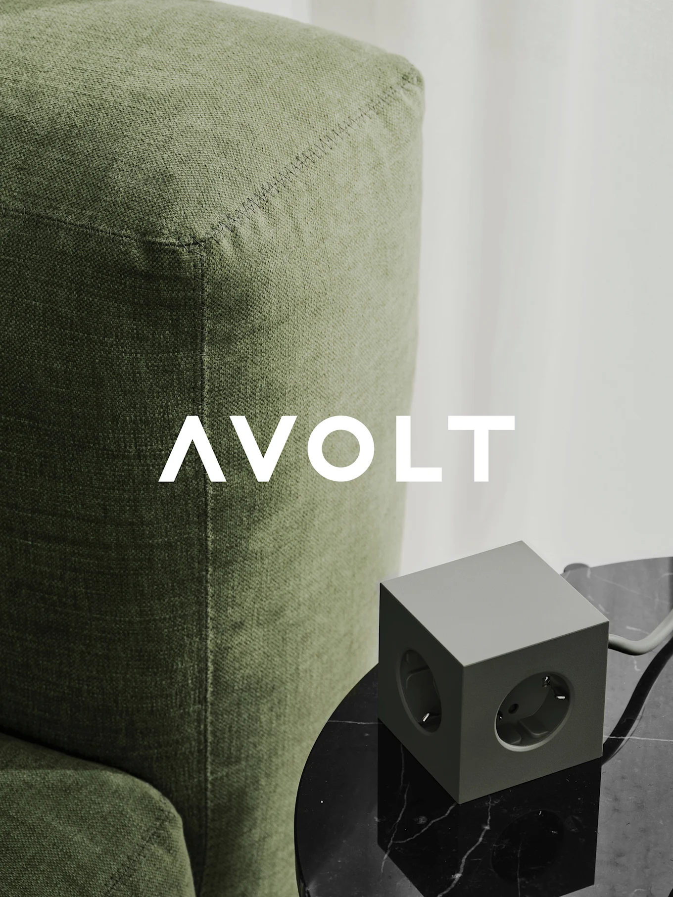 En grønn kubeformet strømskinne med tre uttak på et svart marmorbord ved siden av en grønn sofa. AVOLT-logo.