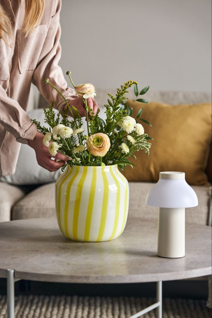En blomsterbukett i fargerik Juice Wide vase i gult og hvitt ved siden av PC portable bordlampe i hvitt fra HAY.
