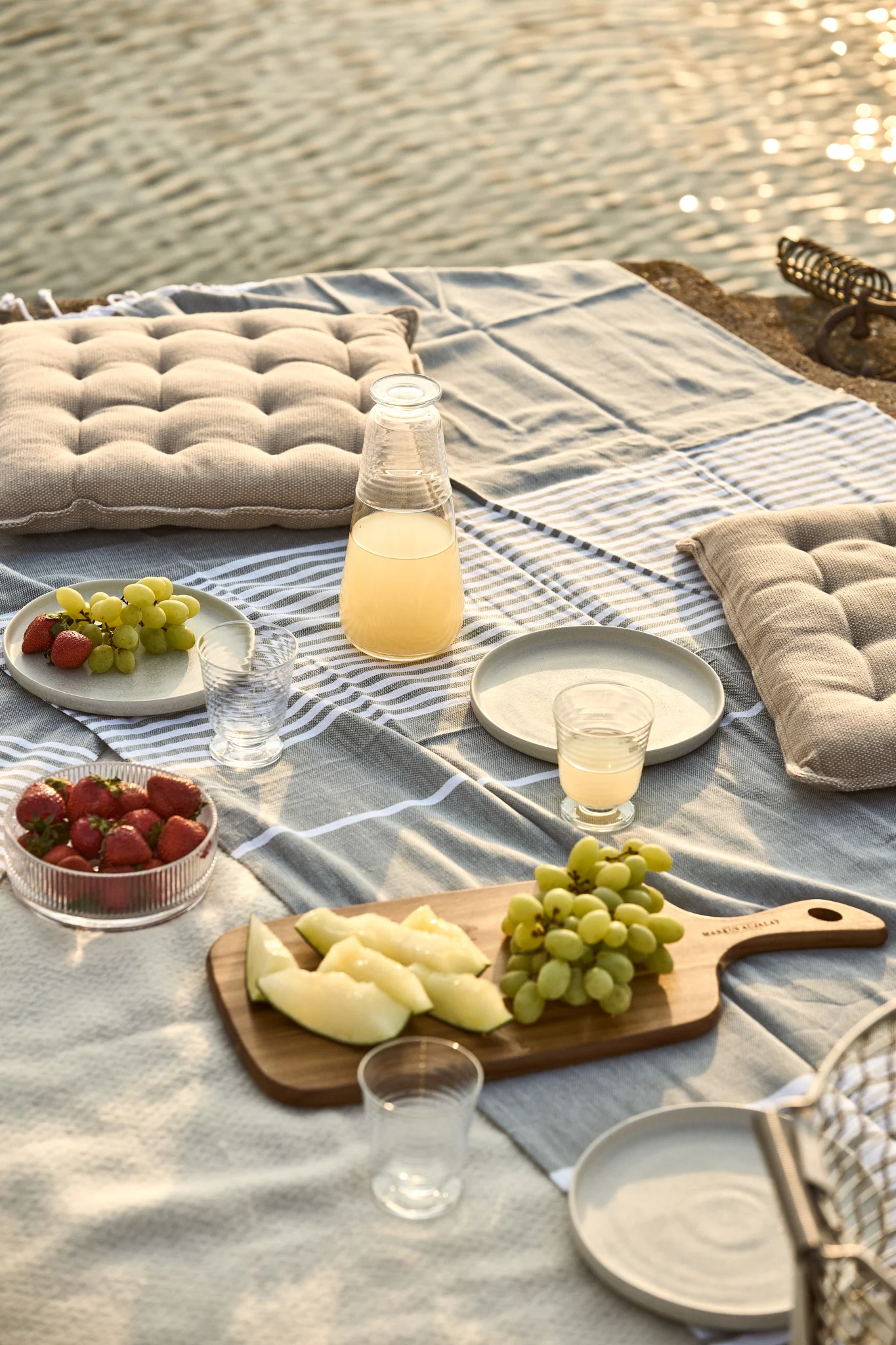 Piknik ved vannet med stripet teppe, puter, juice, jordbær, druer og melonskiver.