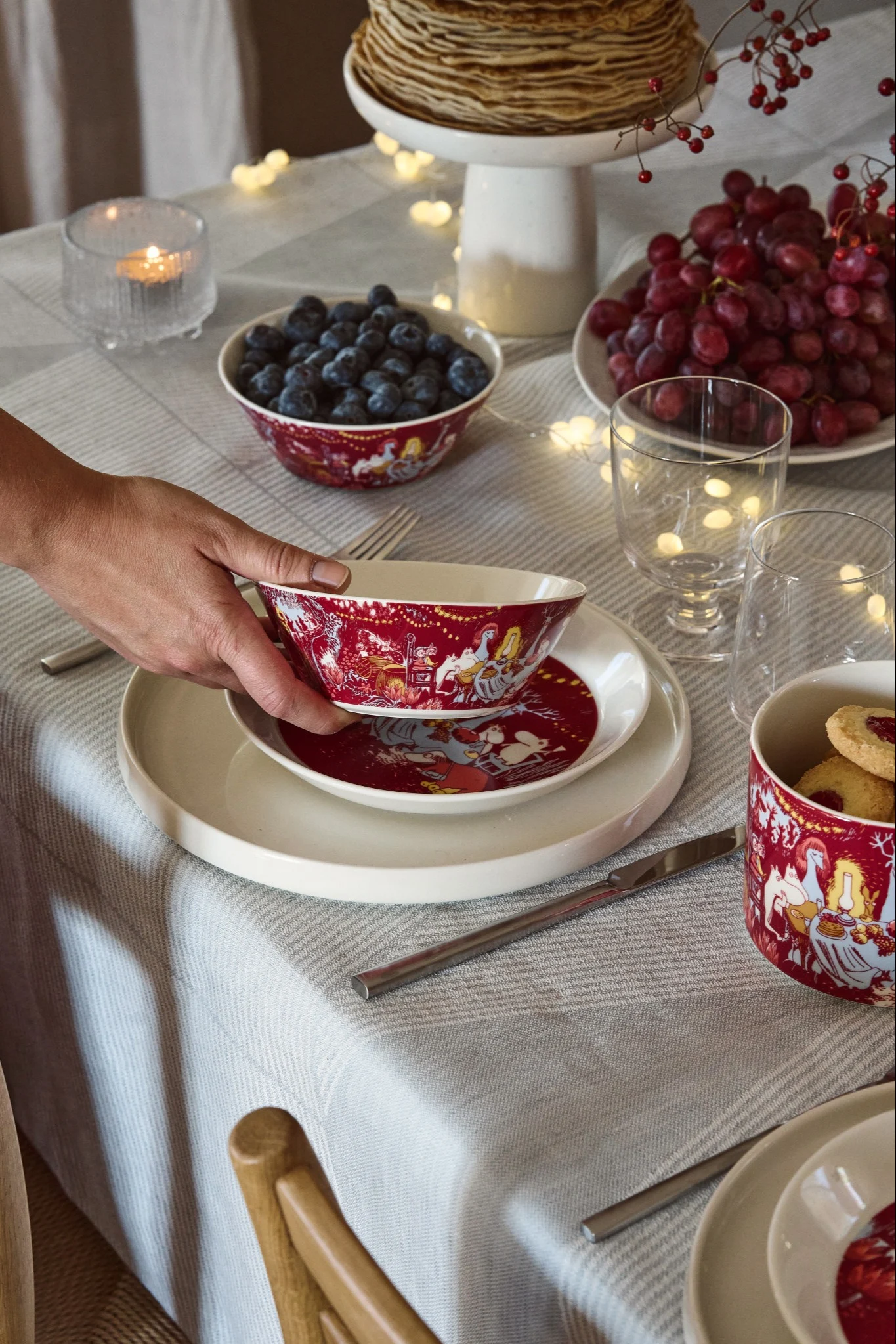 En leken borddekking jul 2025 skapes med rødt servise pyntet med Mummi-motiv, kombinert med fargerike bær som gir en varm og innbydende stemning på bordet.
