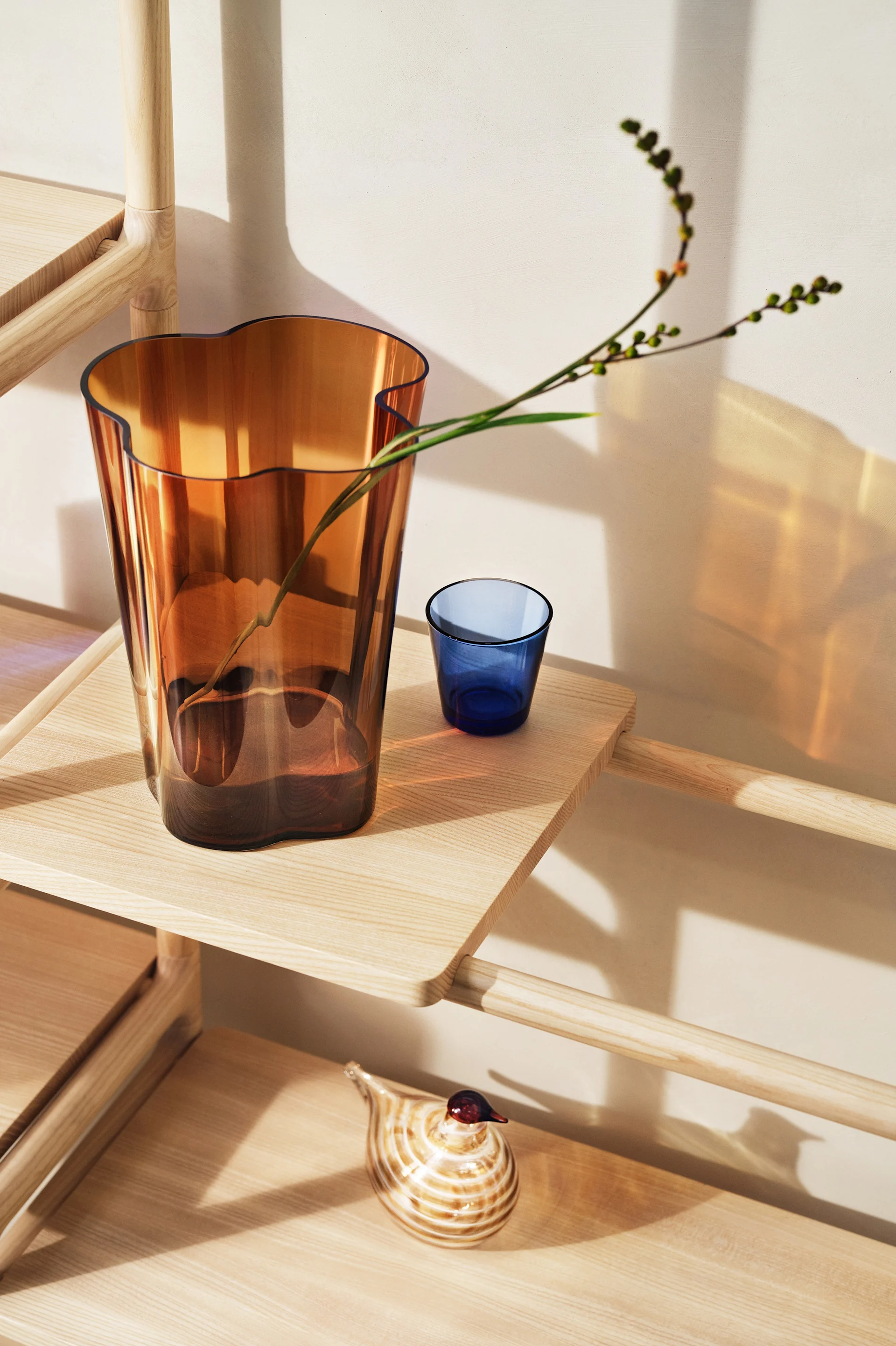 Ravfarget glassvase med bølget topp og gren, blått glassglass og stripet glassfugl på en lys trehylleenhet.