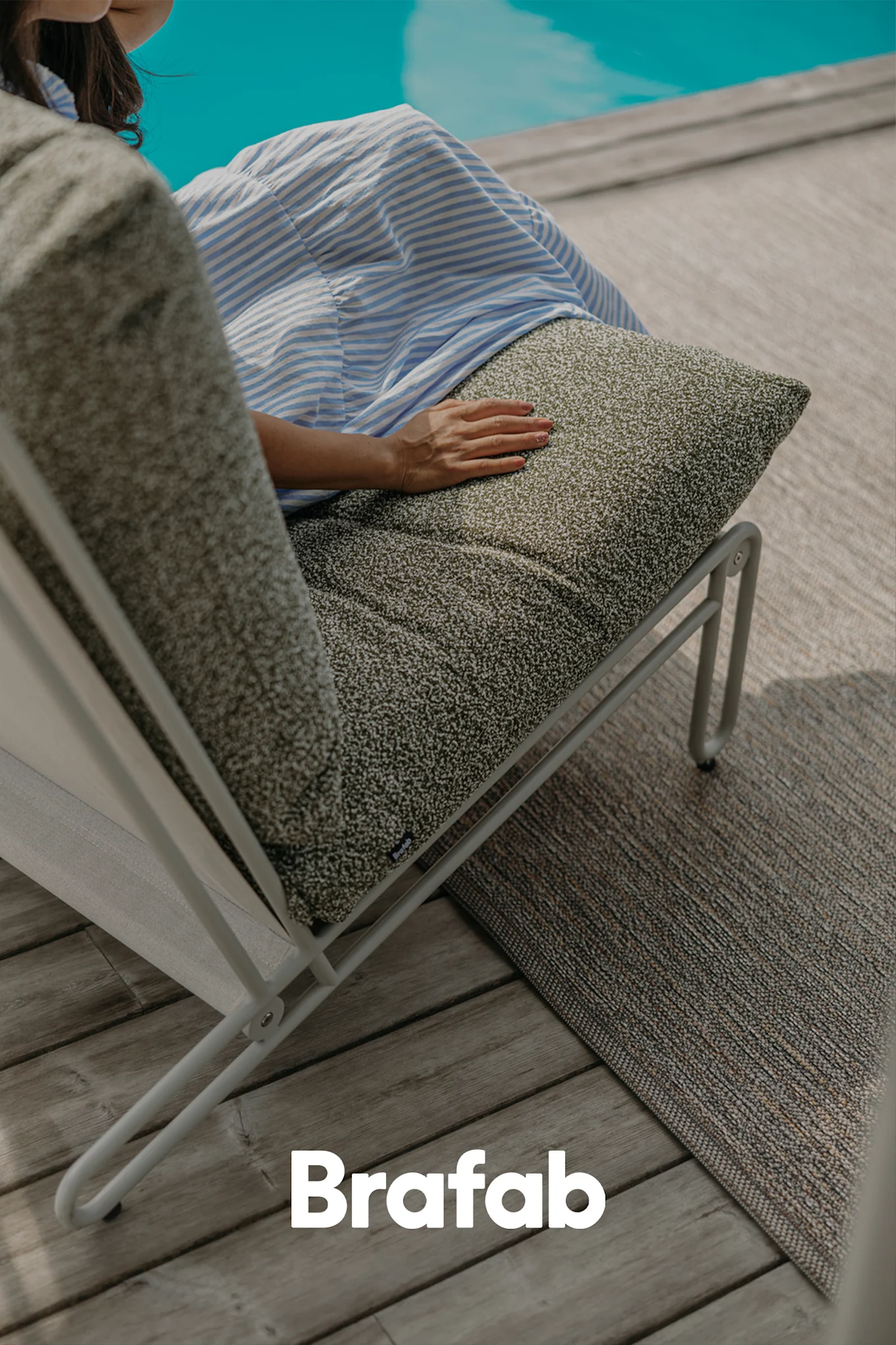 Person sitter på en moderne utendørs solseng med mørkegrønn spraglete pute på et tredekk ved et basseng.