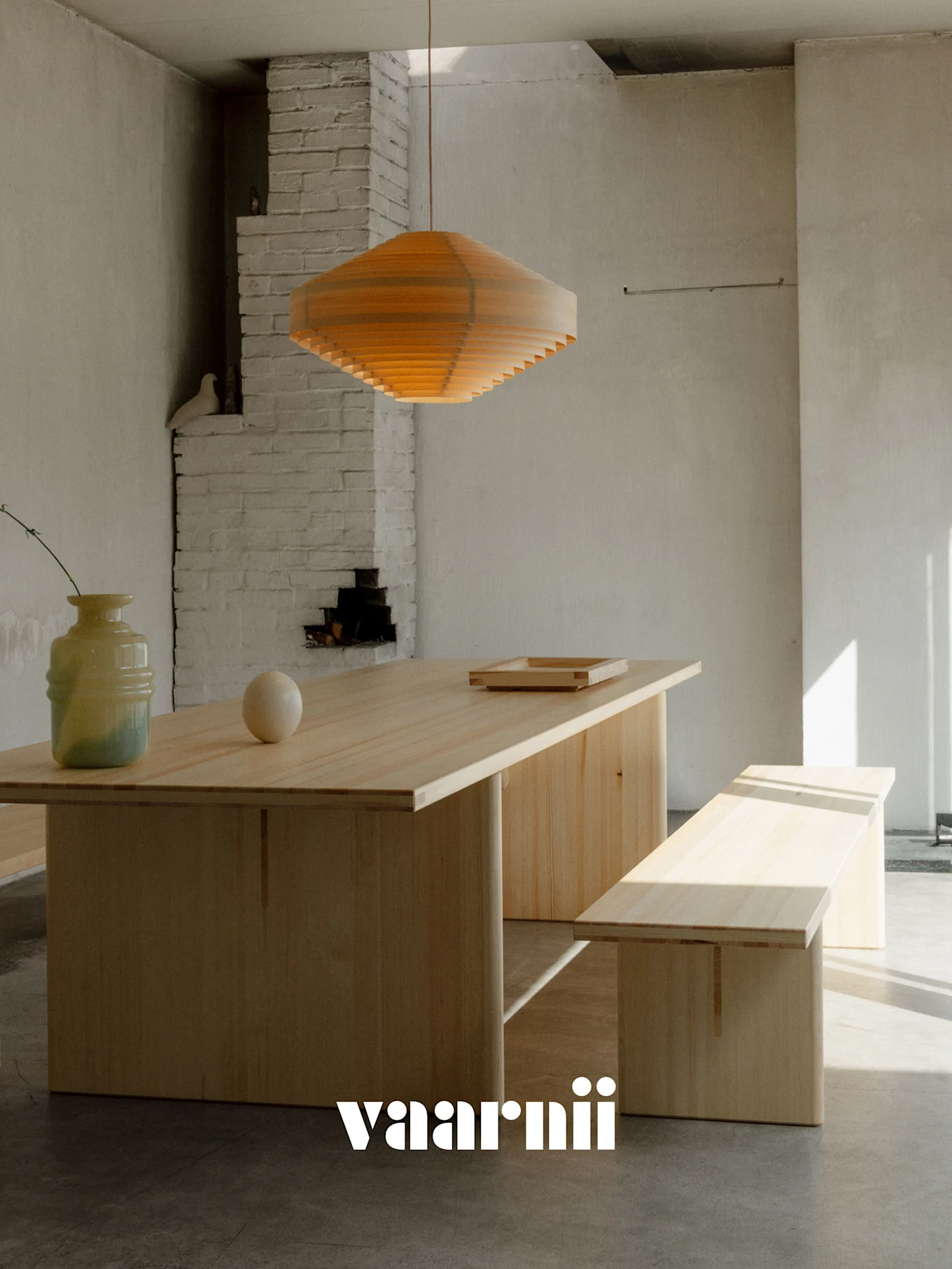 En minimalistisk spisestue med et langt lyst trebord, matchende benk, lagdelt trependellampe, vase og kule. Vaarnii-logo.