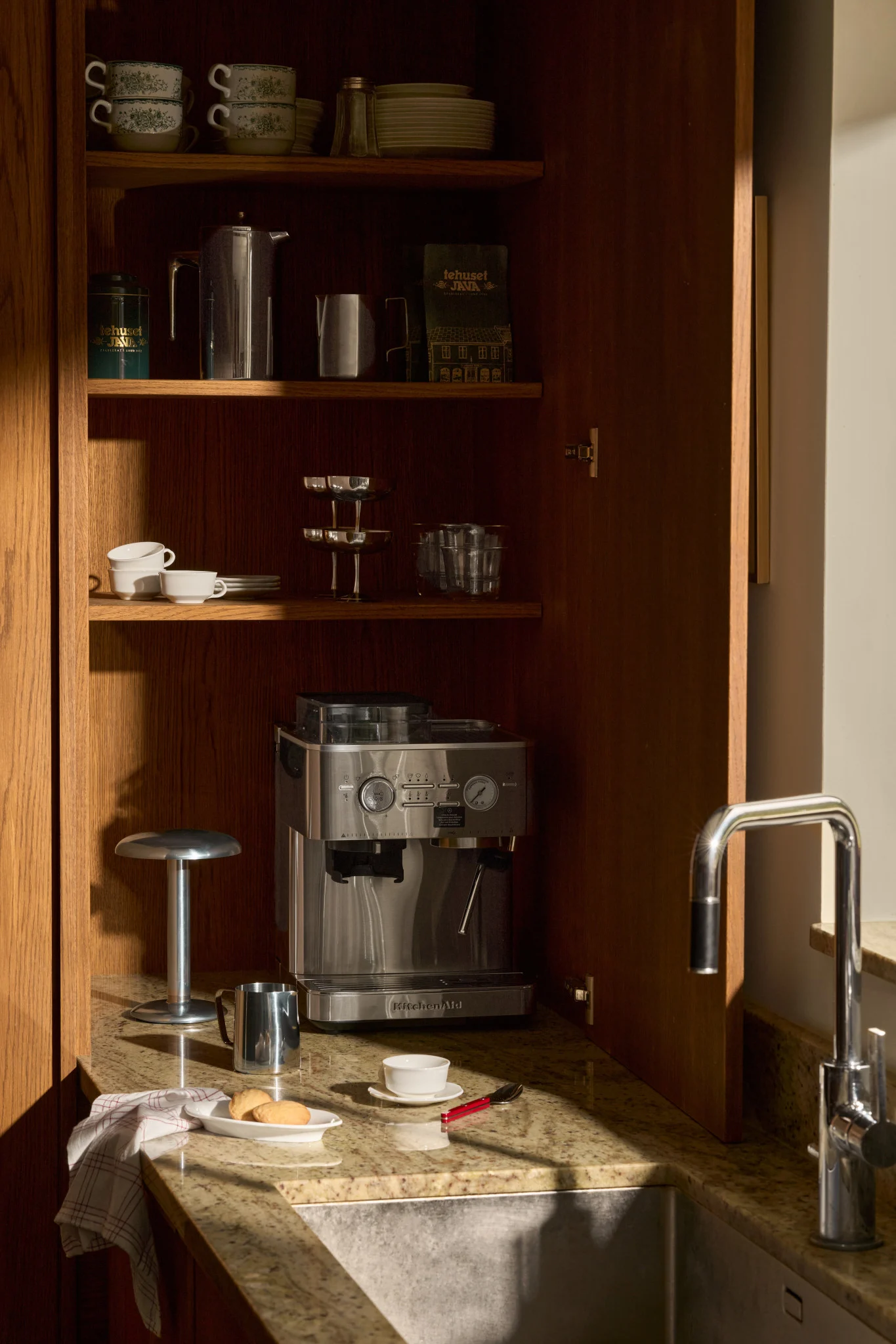 Et åpent kjøkkenskap i tre med espressomaskin, kaffetilbehør, stablede krus og glass på hyller, ved siden av en vask.
