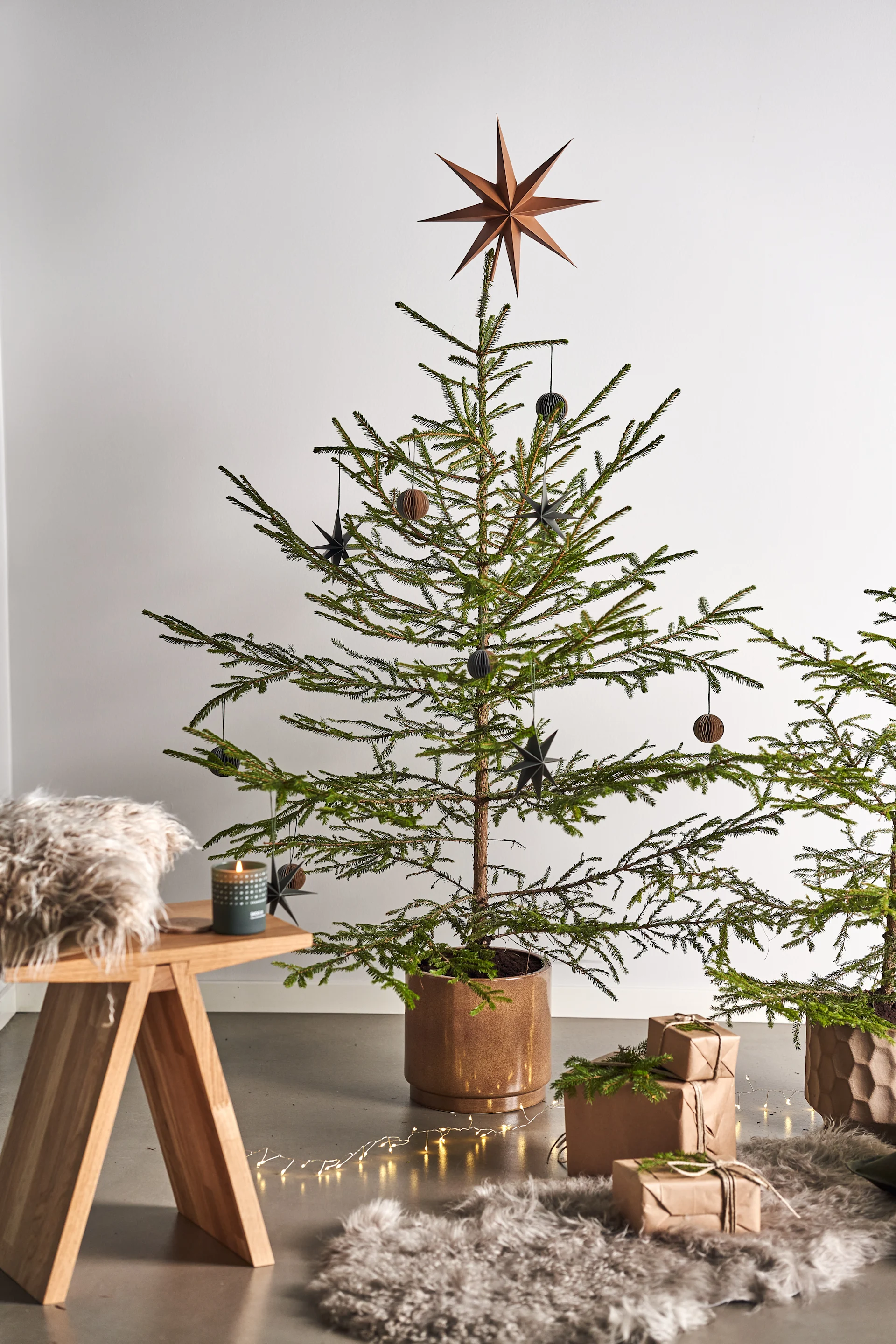 Pynt juletreet med juletrepynt i 4 forskjellige stiler i henhold til Nest Trends - Nurture, Share, Boost and Cultivate. Her ser du Angle krakk fra Muubs med et Skandinavisk duftlys i Forest duften ved siden av pakker på et saueskinnsteppe.