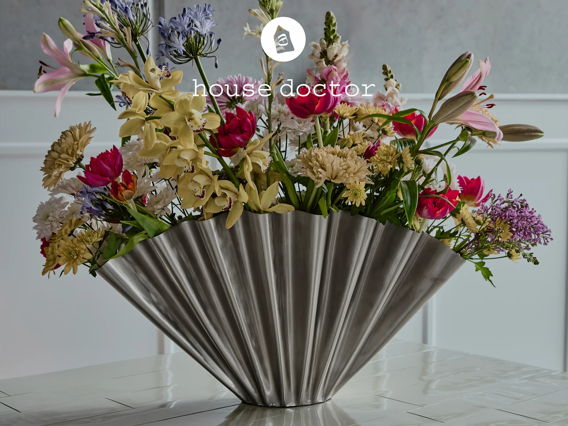Fargerik bukett i riflet sølvvase: rosa liljer, gule orkideer, blå blomster, røde tulipaner. 'house doctor'-logo.
