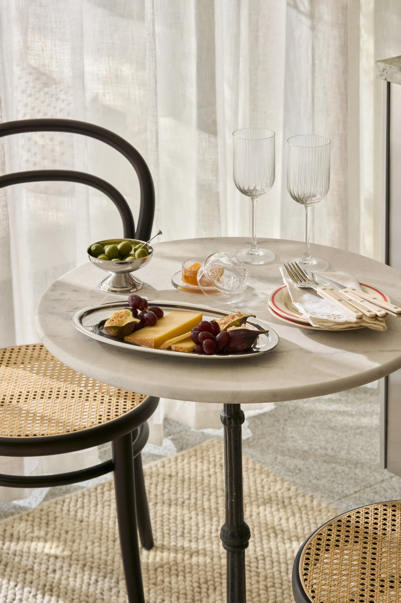Et marmorbord dekket med ost, fiken, druer, oliven, syltetøy, glass og bestikk, med rottingstoler.