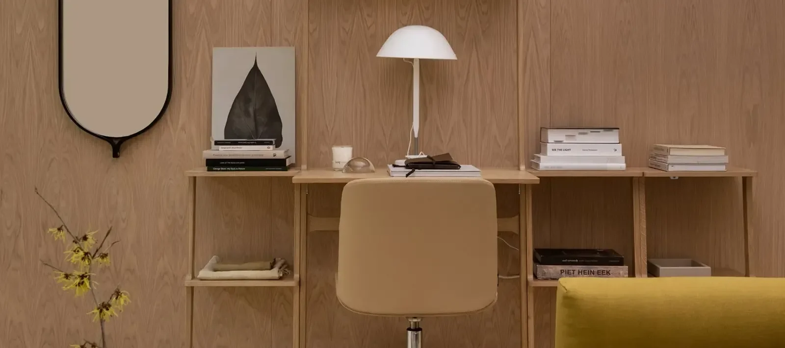 Et moderne hjemmekontor med et lyst treskrivebord, beige stol, hvit lampe og bøker. Et svart speil og gule blomster er synlige.