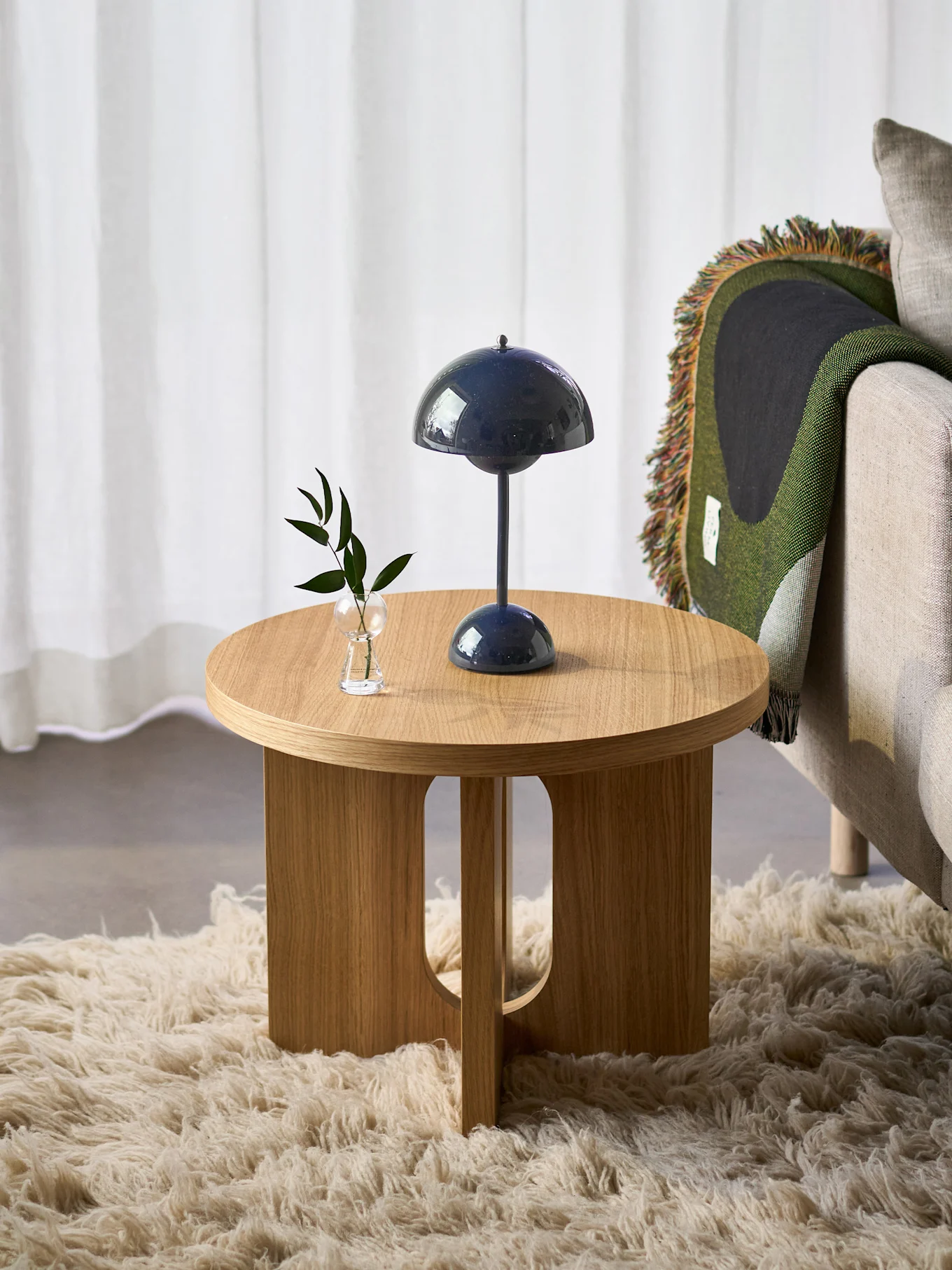 Rundt sidebord i tre med mørkeblå sopplampe og liten plante, ved siden av en lenestol med grønt teppe, på et lodden teppe.