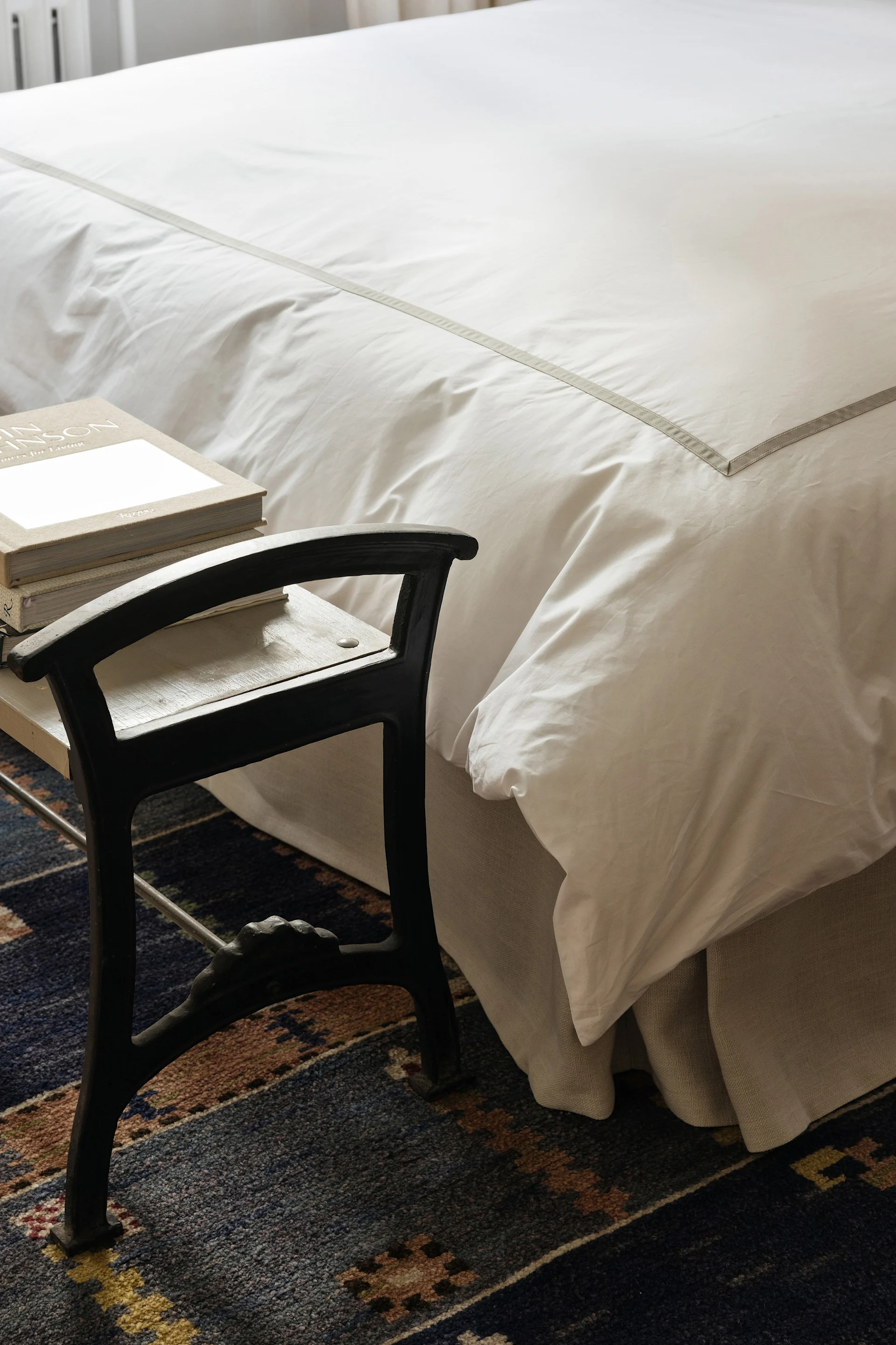 En seng med hvitt sengetøy og en grå stripe ved siden av en mørk krakk med bøker på et mønstret blått teppe.