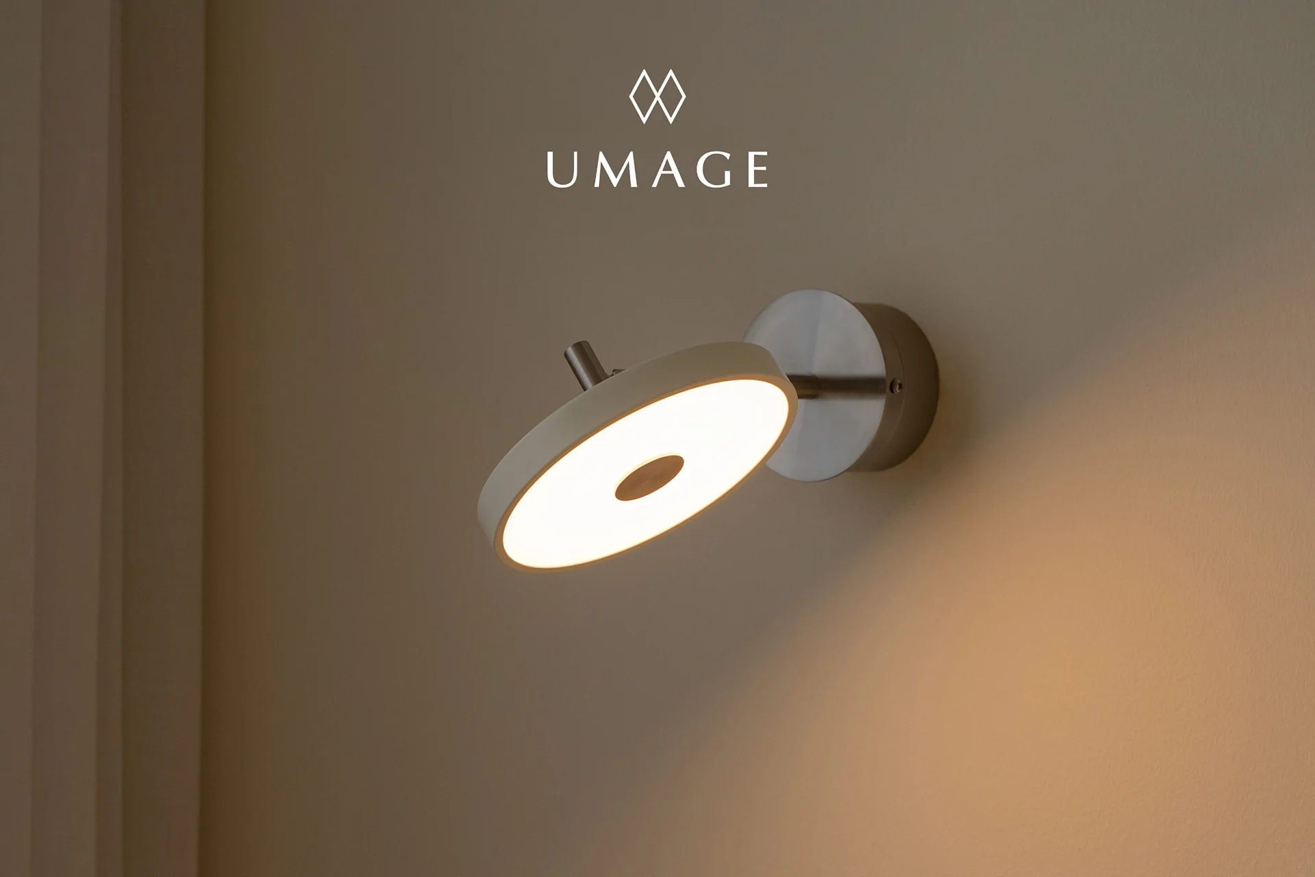 En moderne sirkulær vegglampe med varmt lys, montert på en lysebrun vegg. UMA-logo øverst.