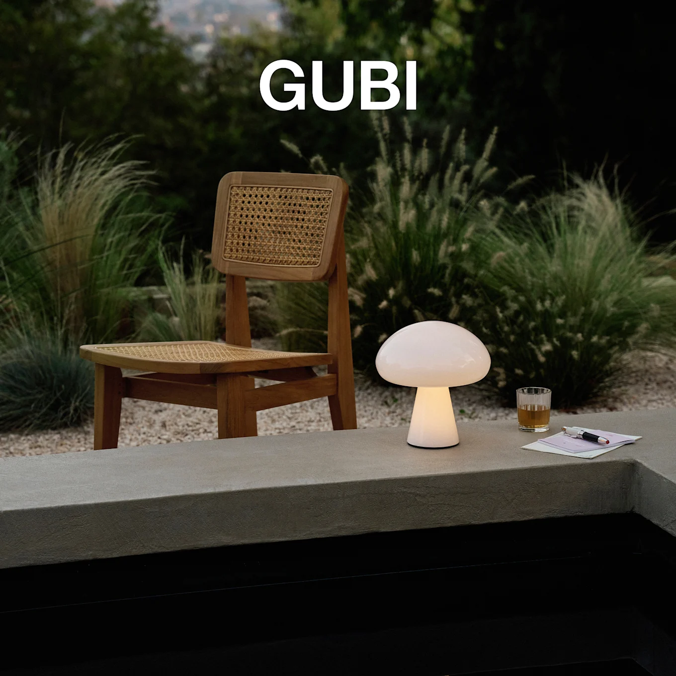 GUBI-logo. En opplyst soppformet bordlampe, et glass og en penn på en betongkant, med en trestol og frodig grønt.