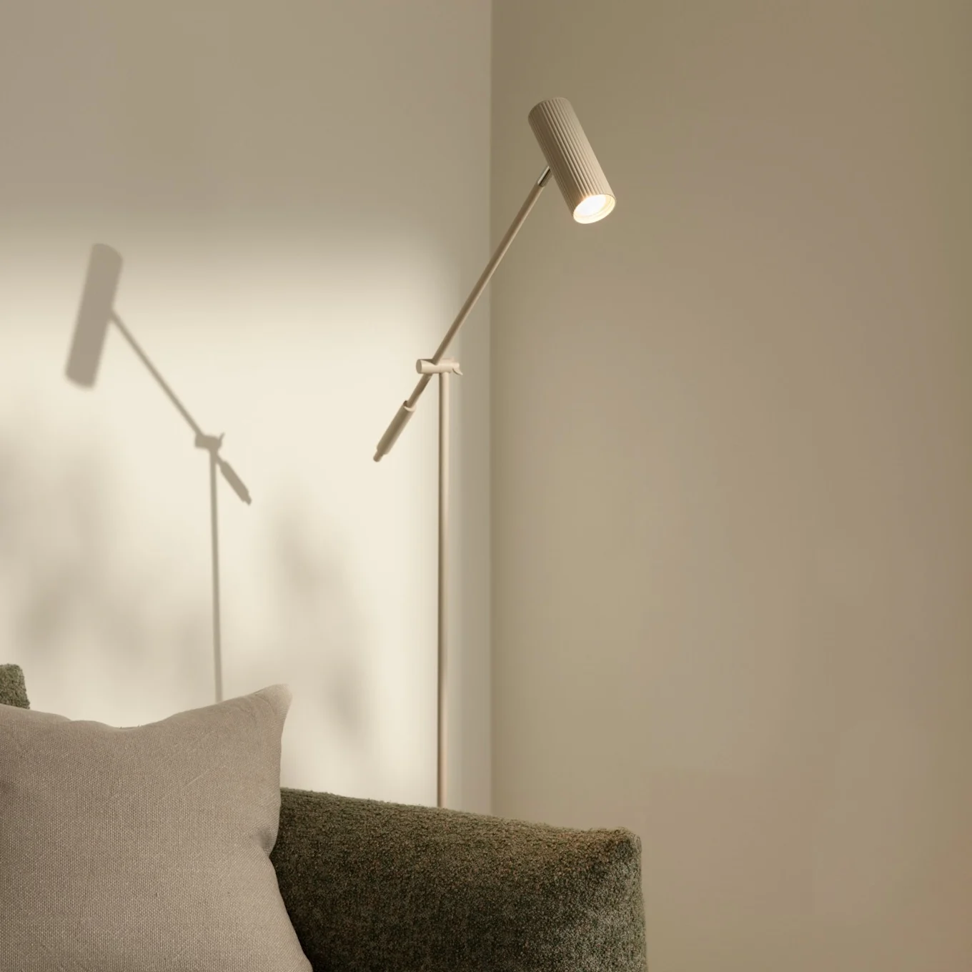 En moderne, slank gulvlampe med ribbet hode som lyser opp en lys vegg, ved siden av en grønn teksturert sofa med en beige pute.