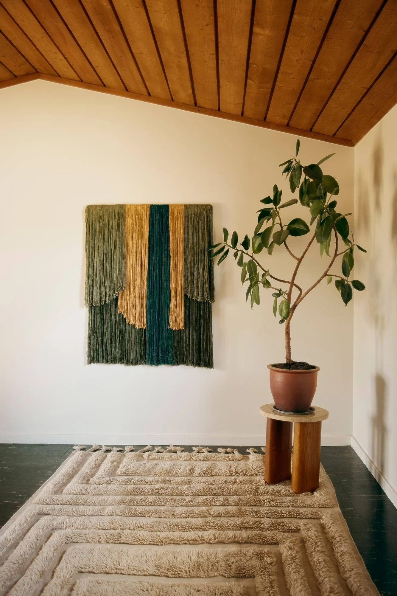 Et rom med et teksturert veggteppe i grønt, gull og teal garn, en potteplante og et raggete beige teppe med geometriske mønstre.